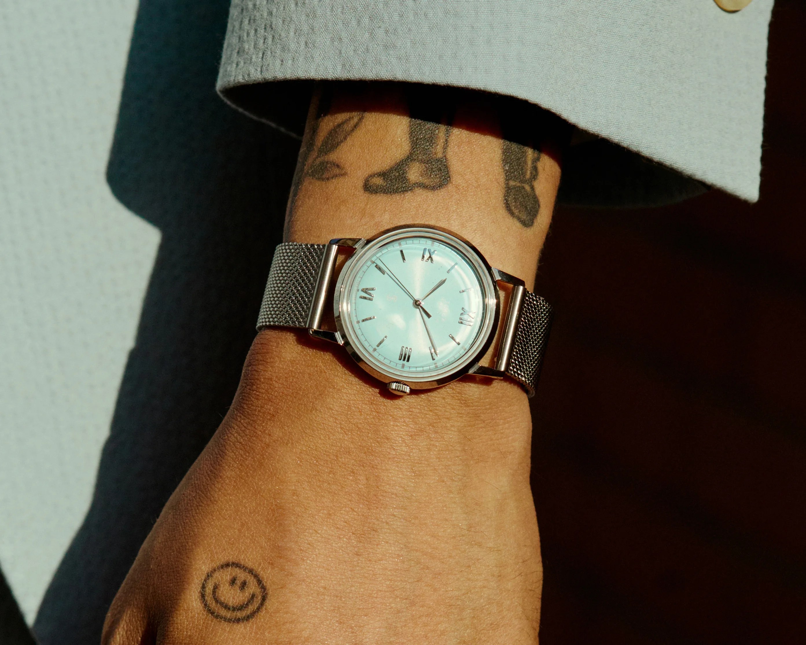 Silver mesh strap wristwatch with a light blue face and Roman numerals worn on a tattooed wrist.
