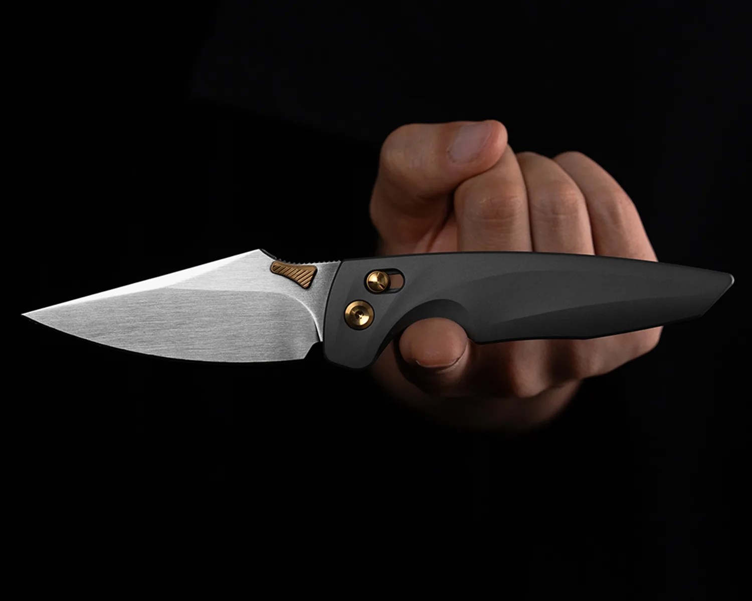 Hand holding a folding knife with a silver blade and black handle with gold screws against a black background.