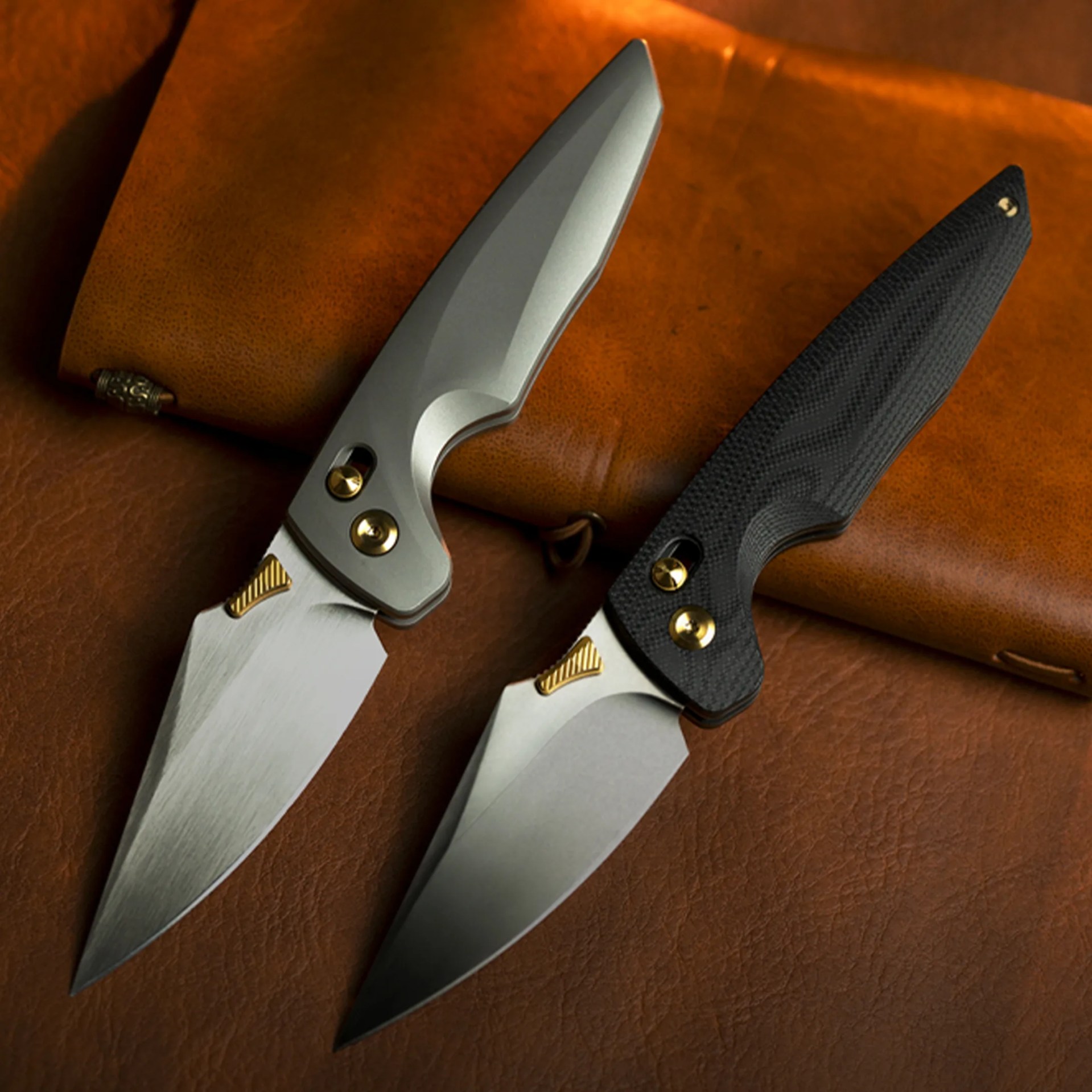 Two folding knives with silver blades and gold accents, one with a silver handle and the other with a textured black handle, on a brown leather surface.