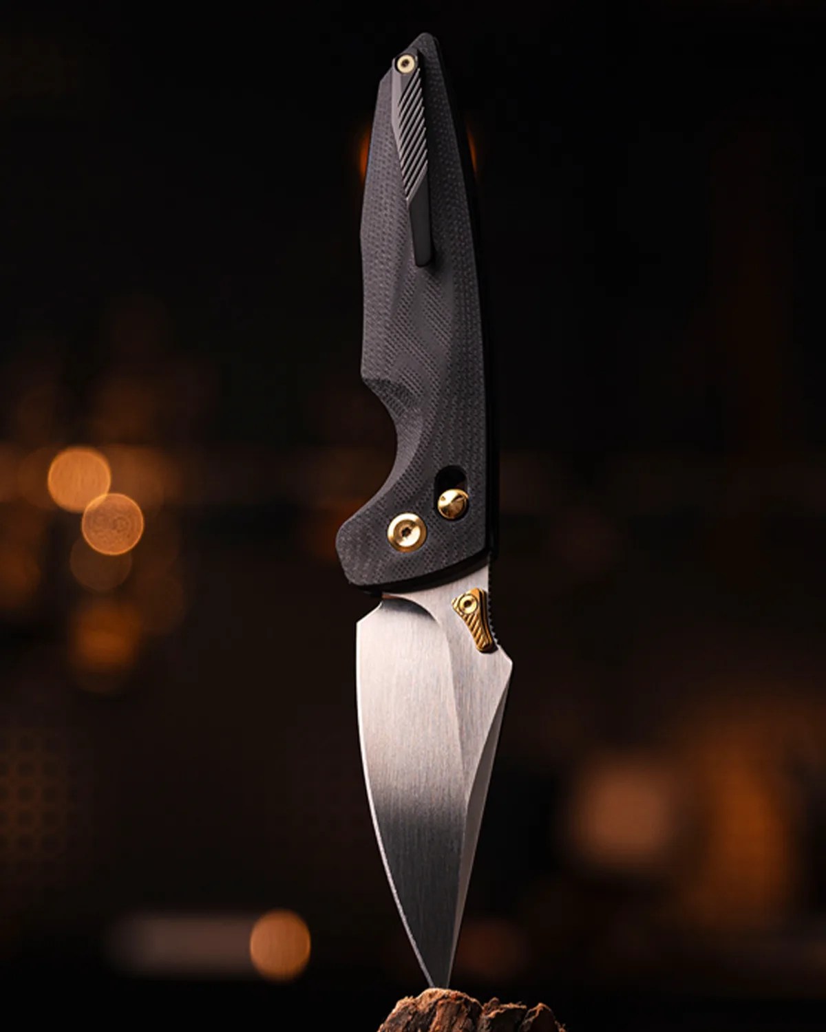 Folding knife with a textured black handle and silver blade with gold accents, standing tip-down on wood.