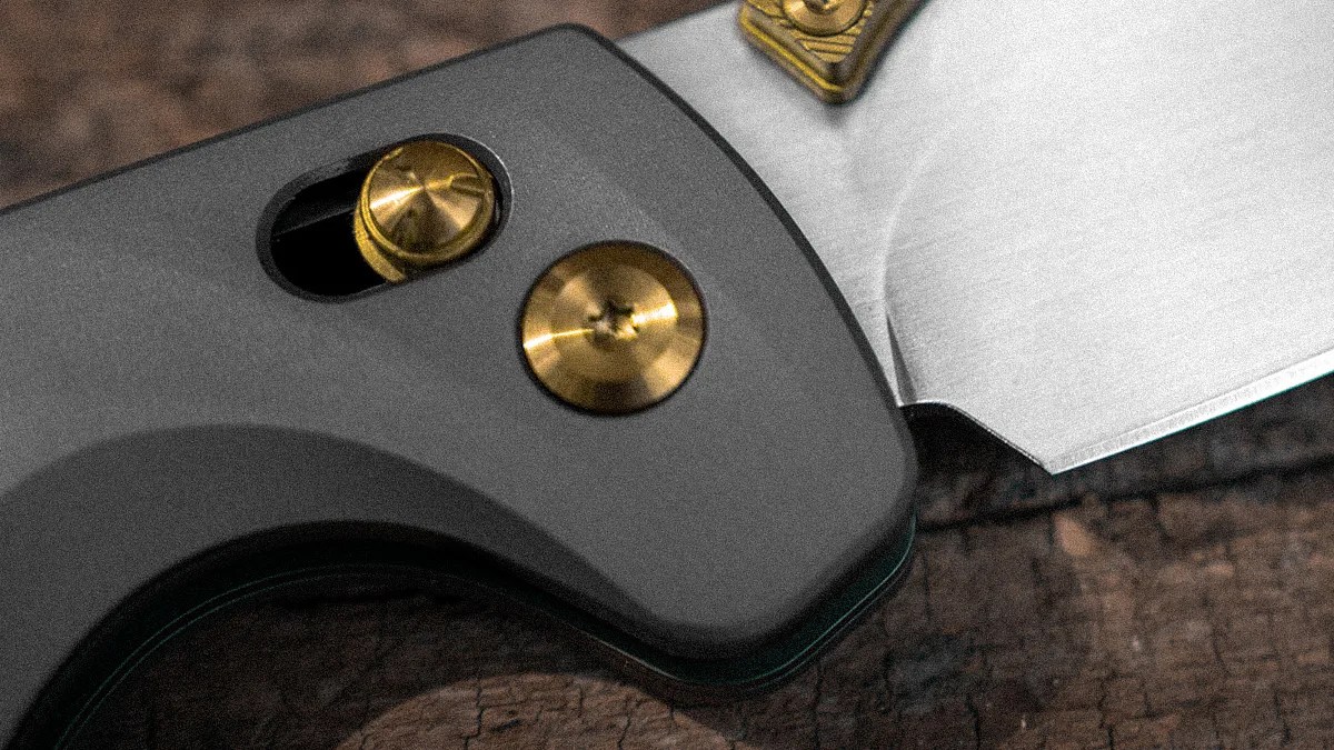 Close-up of a folding knife with a matte black handle and brass pivot screws on a wooden surface.