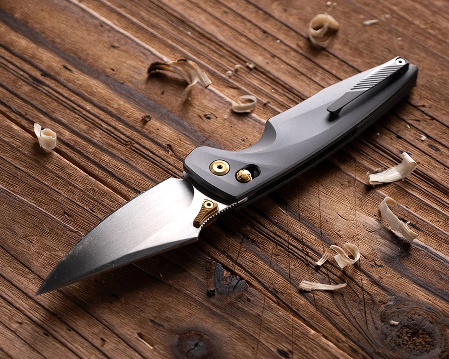 Folding knife with a silver blade and gray handle featuring gold screws, resting on a wooden surface with wood shavings.
