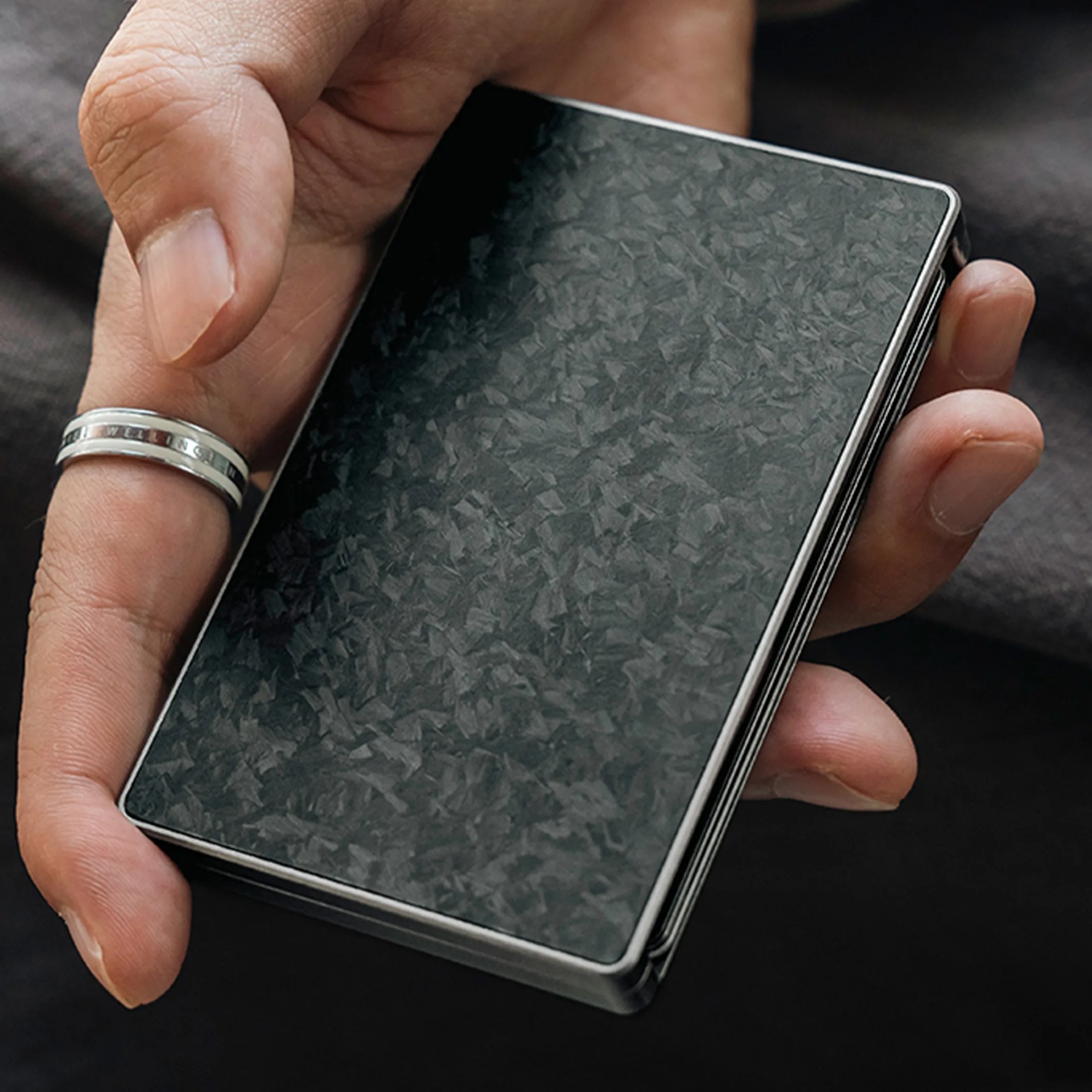 Hand holding a black textured rectangular object with a silver frame and a silver ring on the thumb.
