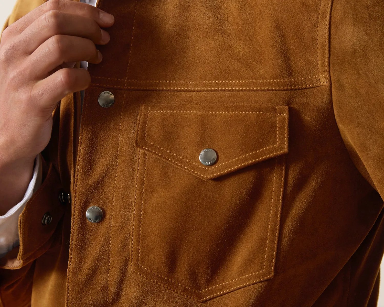 Close-up of a brown suede jacket pocket with silver snap buttons and visible stitching, held by a hand.