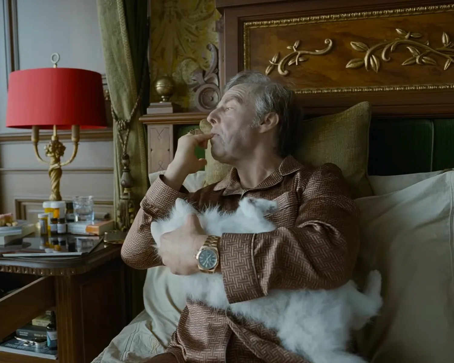 Man in patterned brown pajamas holding a fluffy white cat while sitting on a bed with a carved wooden headboard.