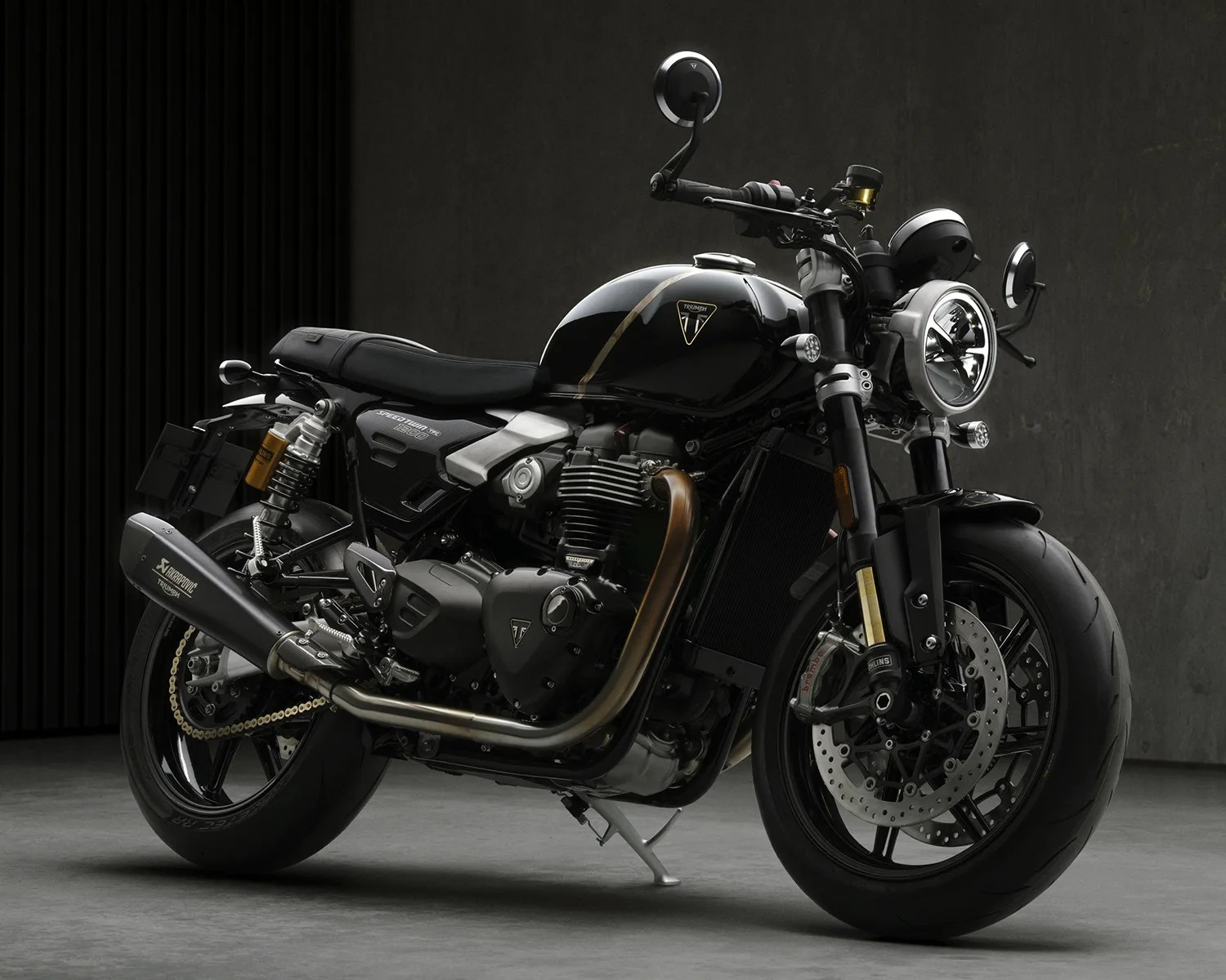 Black Triumph Speed Twin 1200 motorcycle with silver exhaust and gold suspension in a dark room.