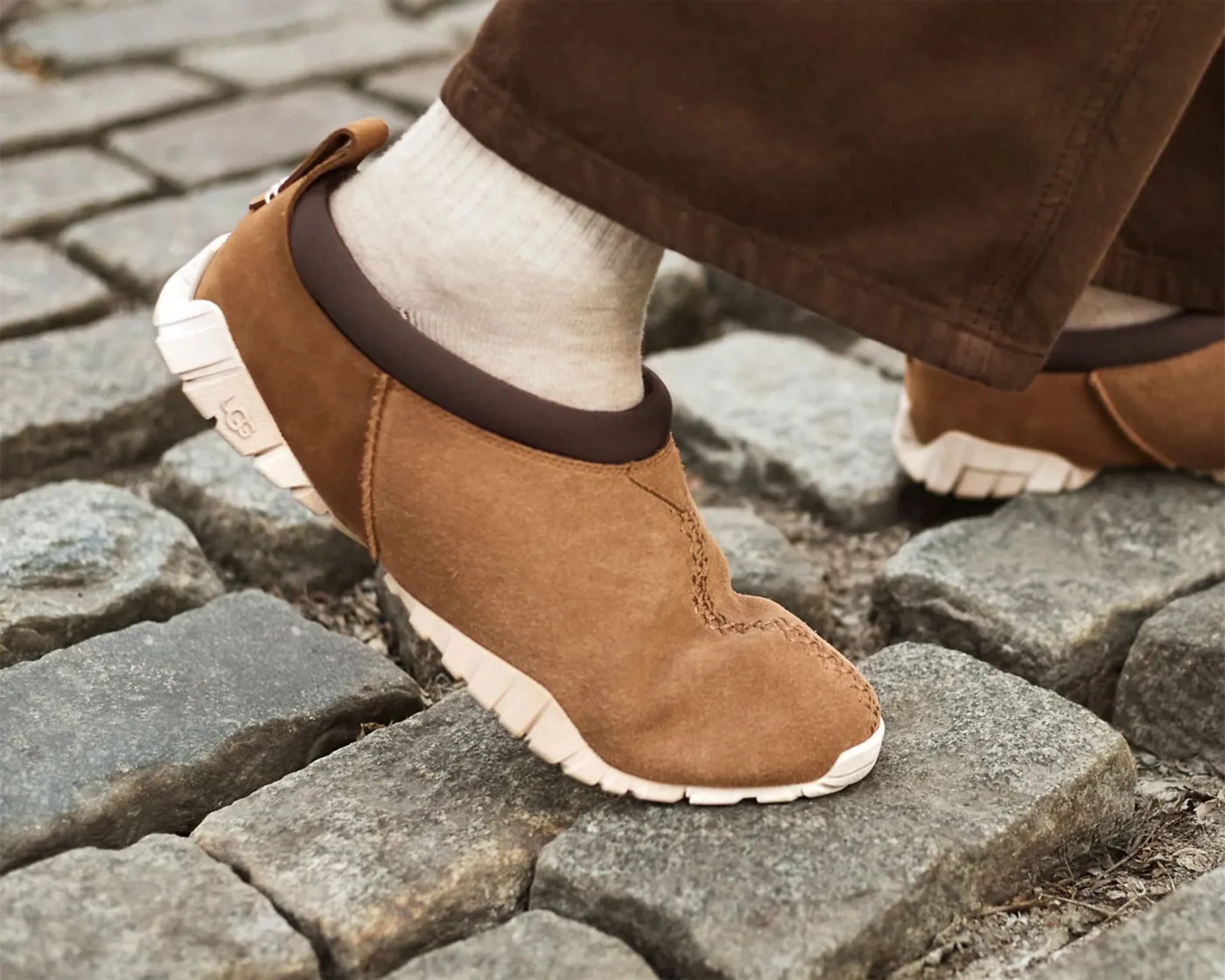 Brown suede slip-on shoes with white soles worn with white socks on cobblestone pavement.