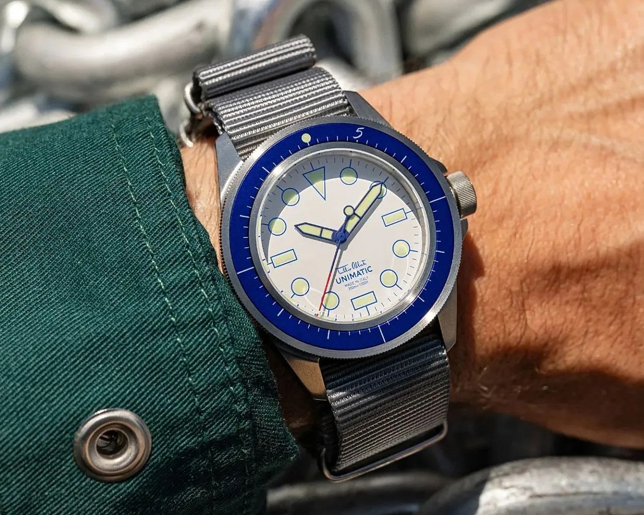 Wristwatch with white dial, blue bezel, luminous hands, and gray fabric strap worn on a wrist.