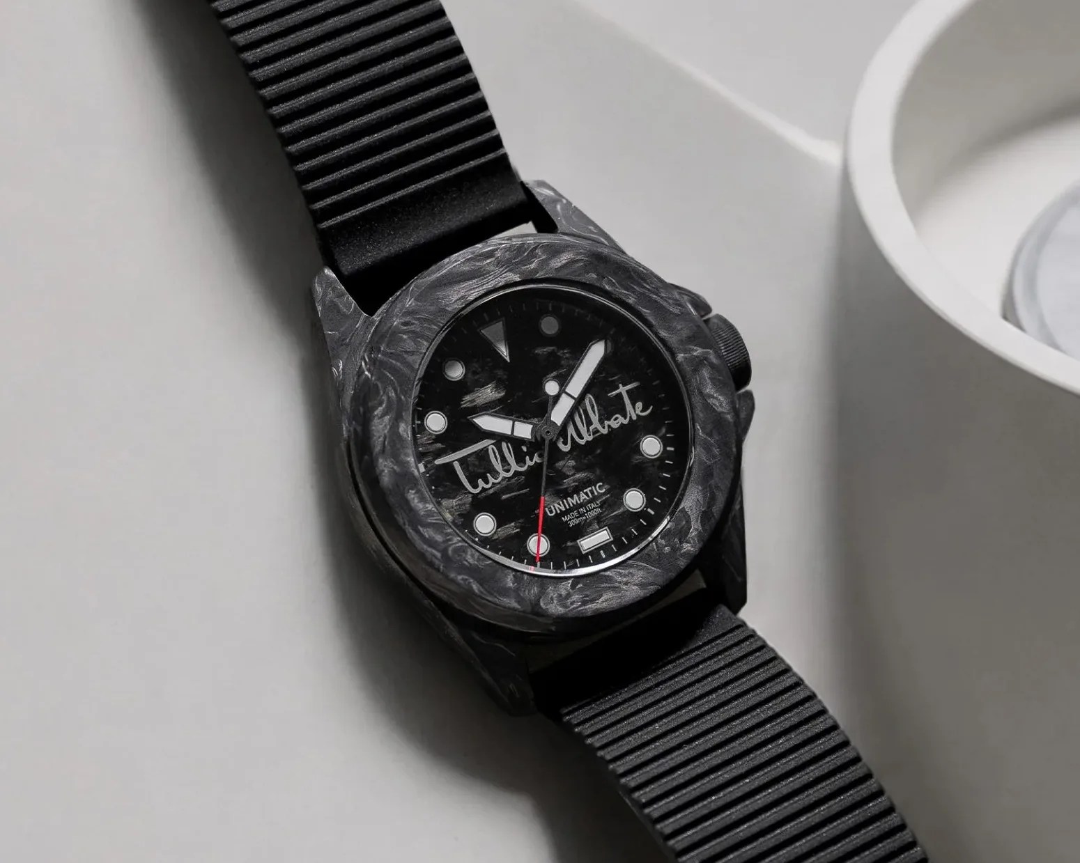 Black wristwatch with marbled bezel, black dial featuring white markers and "Tullio Abbate" signature, on a ribbed black strap.