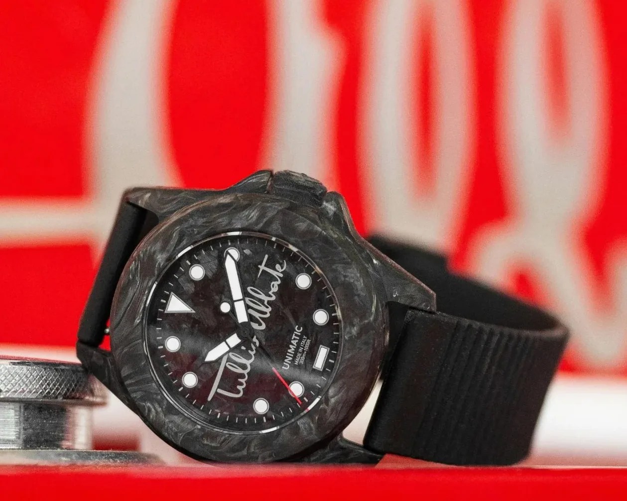 Black marbled watch with white markers and "Tullio Abbate" signature on the dial, black rubber strap, on a red surface.