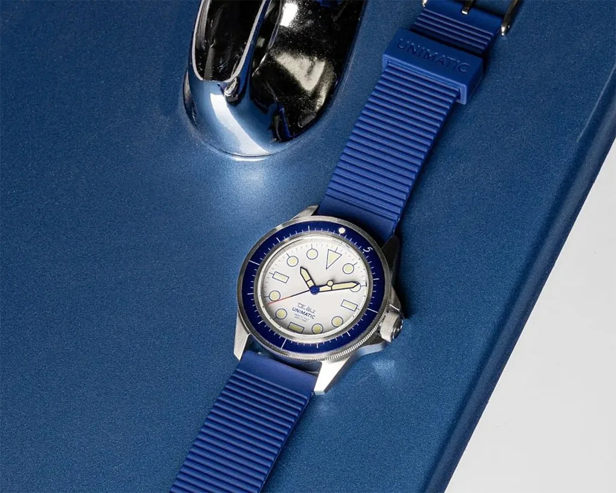 Blue Unimatic wristwatch with white dial and blue bezel on a blue metallic surface.