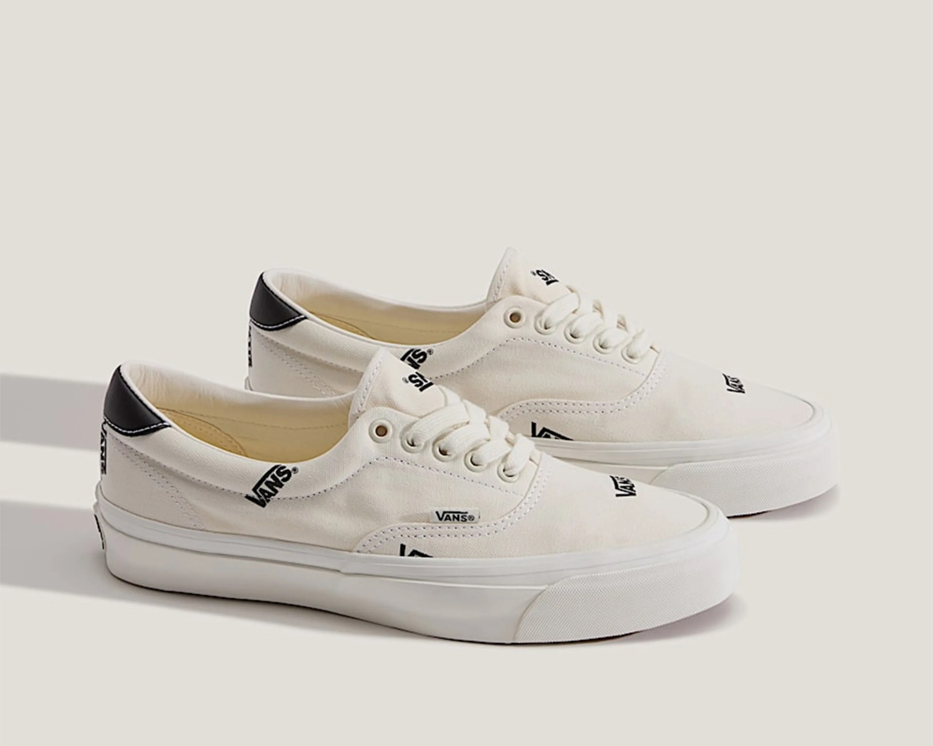 Off-white Vans sneakers with black logo prints and black heel accents on a light background.