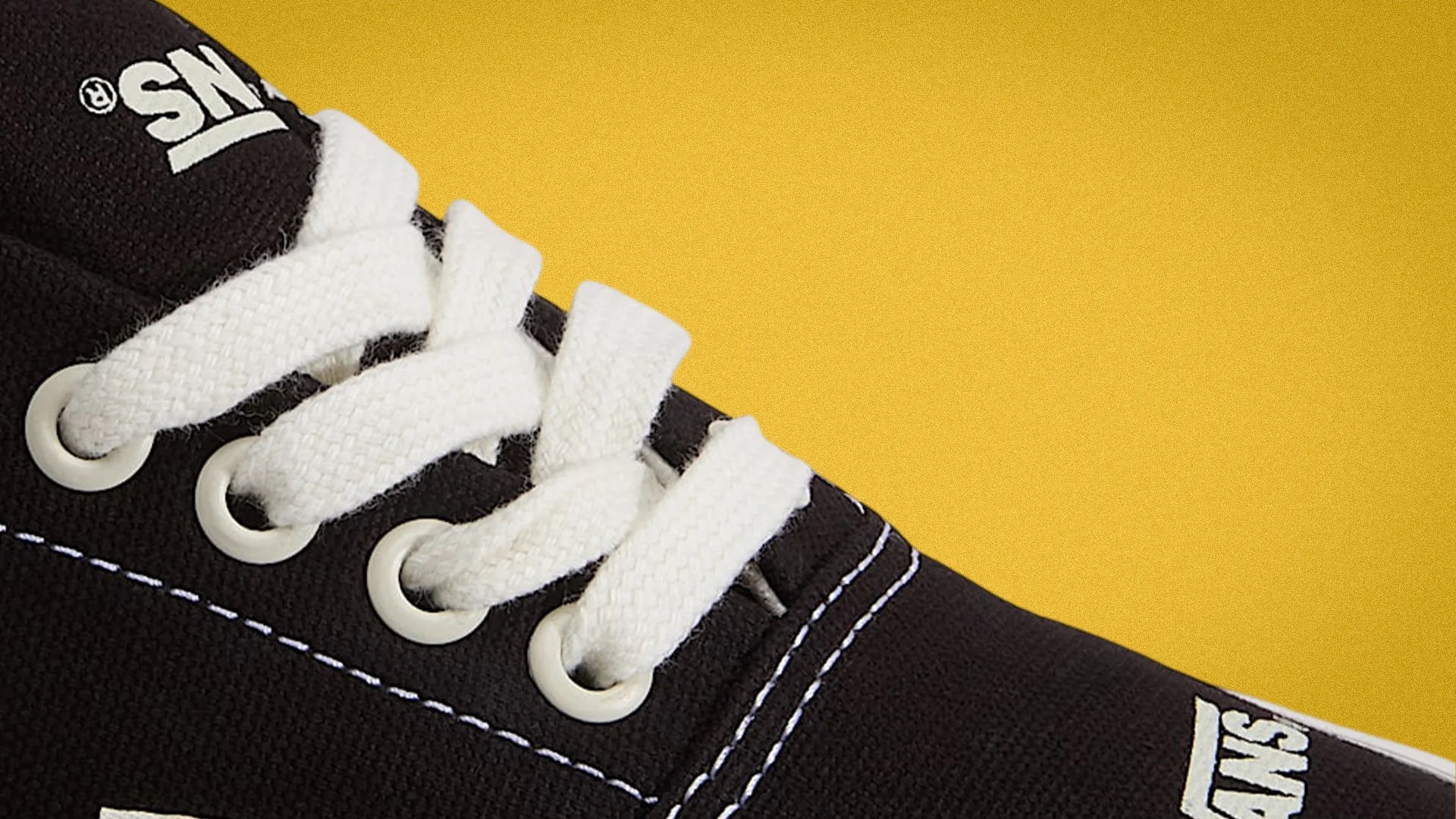 Close-up of black Vans sneaker with white laces and white stitching on a yellow background.