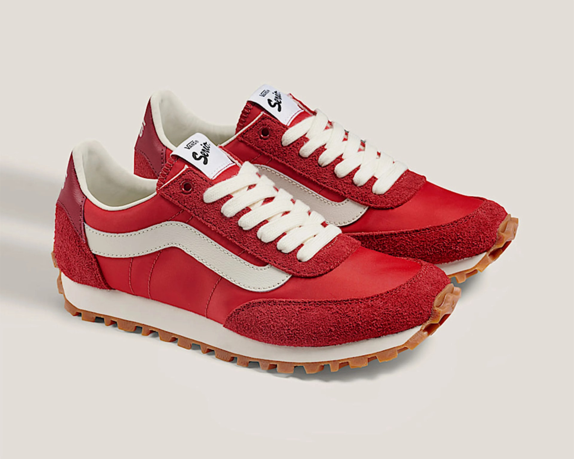 Pair of red Vans sneakers with white side stripe, white laces, and gum rubber soles on a beige background.