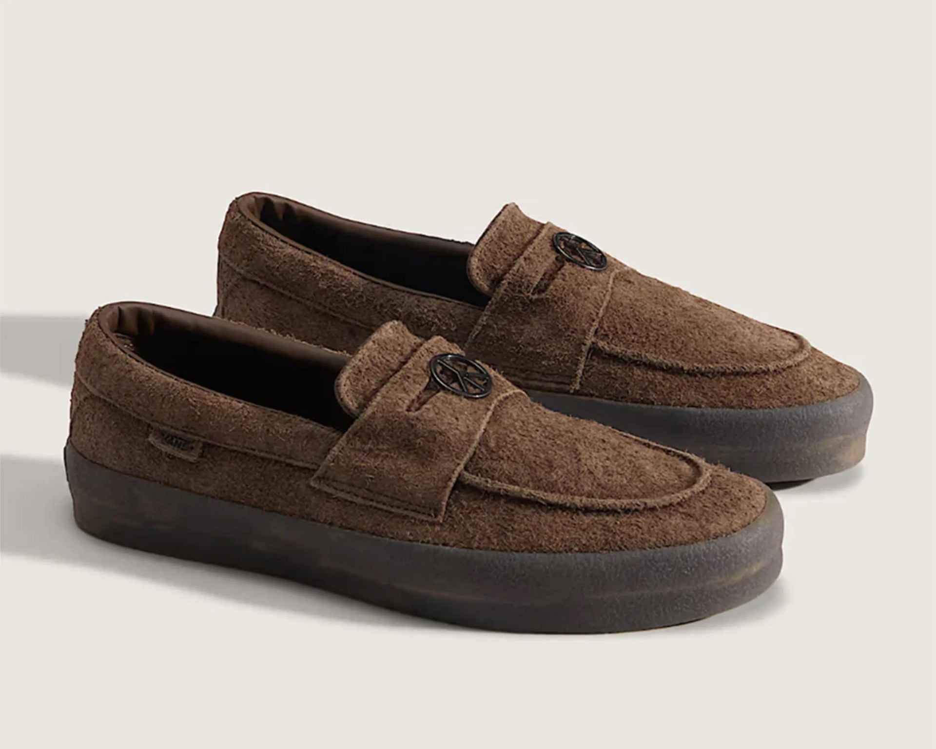 Brown suede loafers with black rubber soles and a round black emblem on the strap.