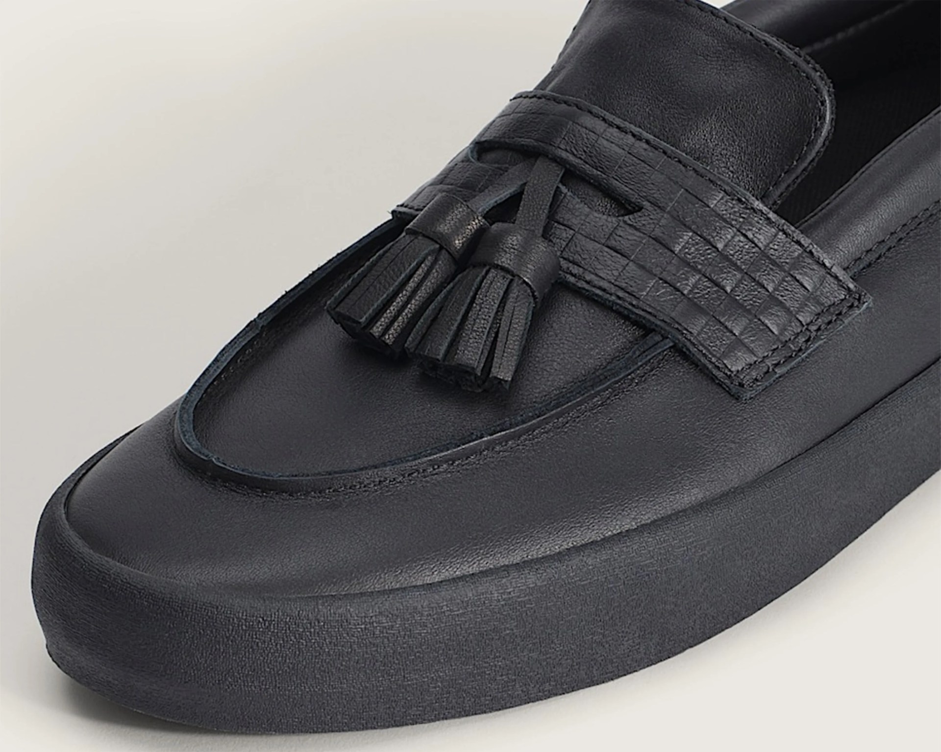 Black leather loafer with tassel detail and textured strap on a light background.