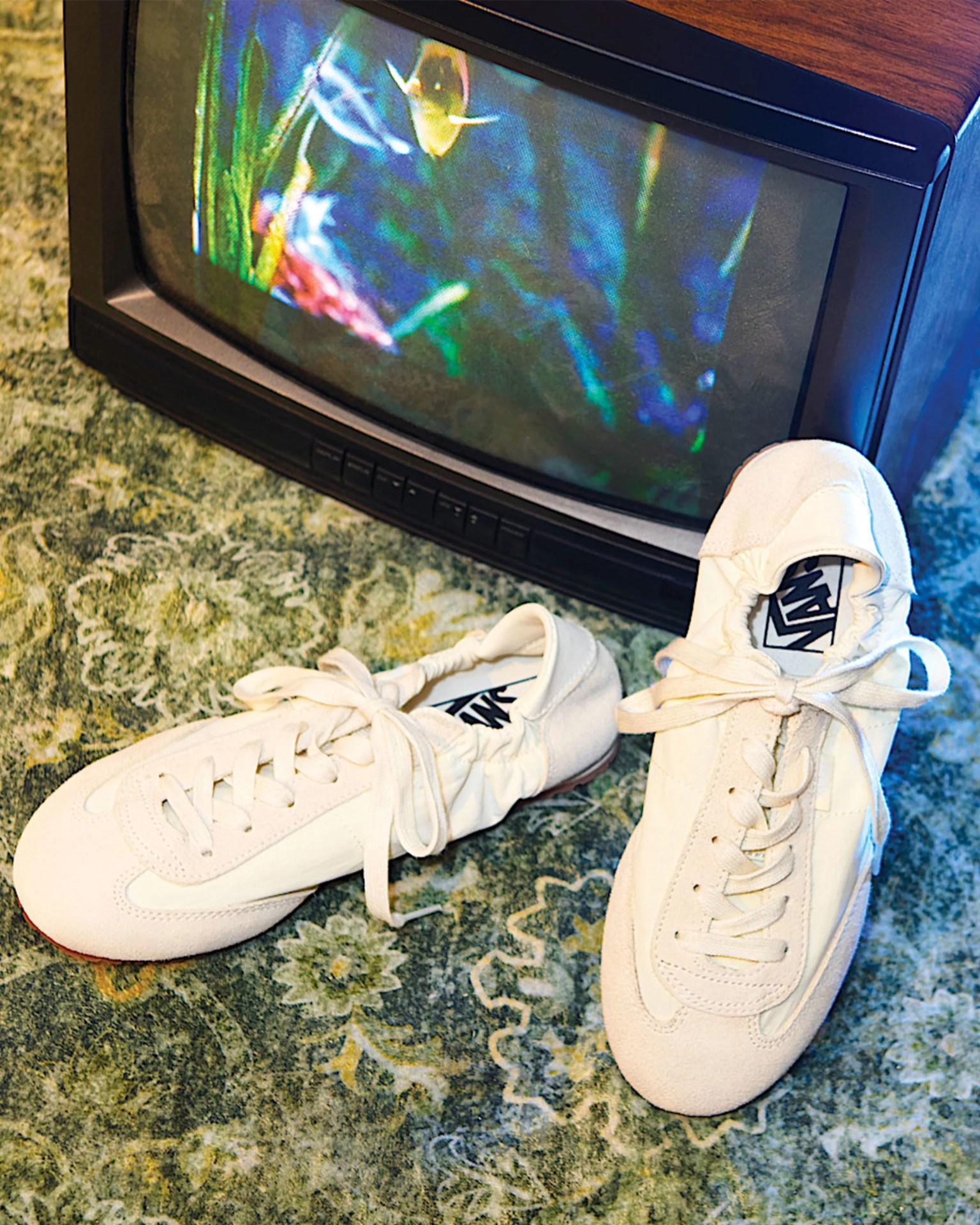 Pair of white Vans sneakers with laces on a patterned green carpet next to a vintage TV showing an aquarium scene.