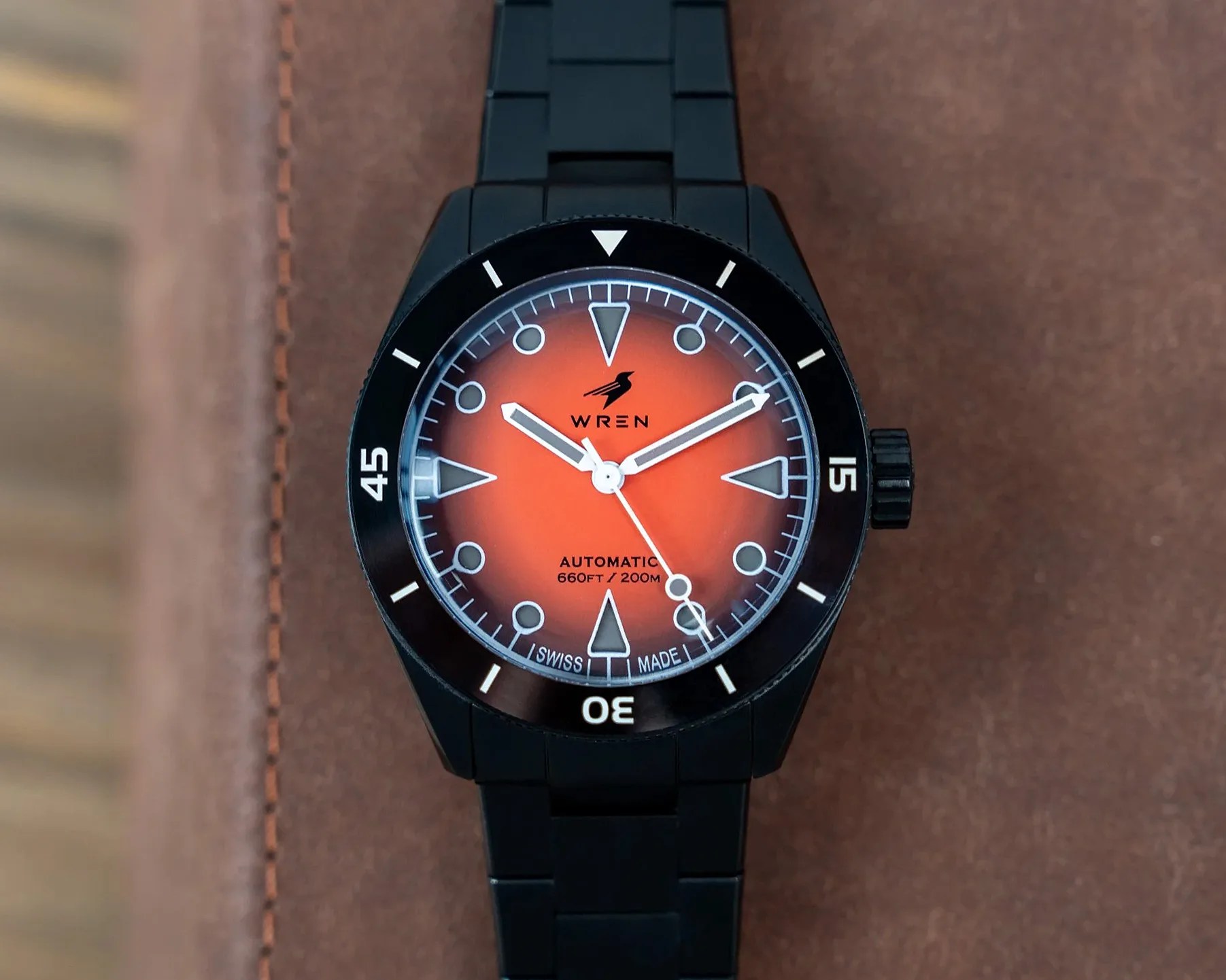 Black metal wristwatch with orange gradient dial and white markers on a brown leather surface.