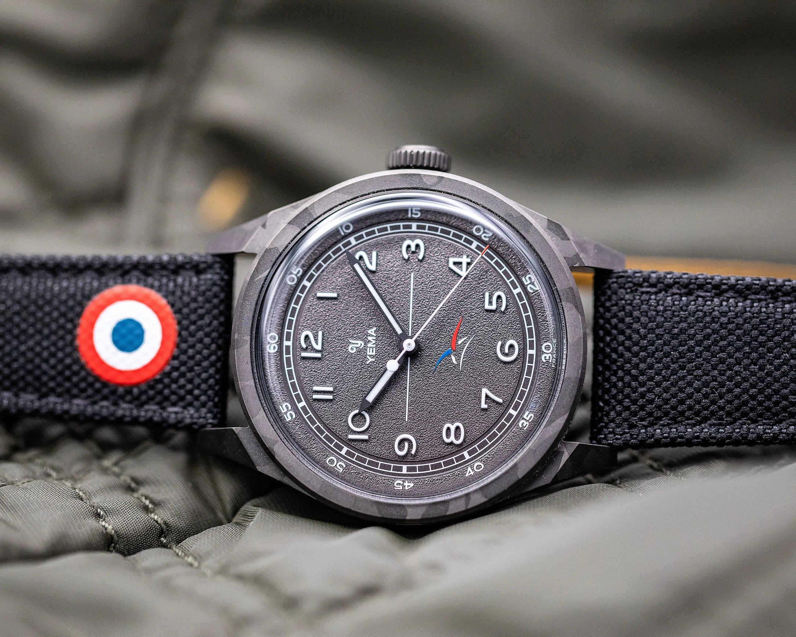 Close-up of a Yema wristwatch with a textured black dial, white numerals, and a black fabric strap with a red, white, and blue circular patch.