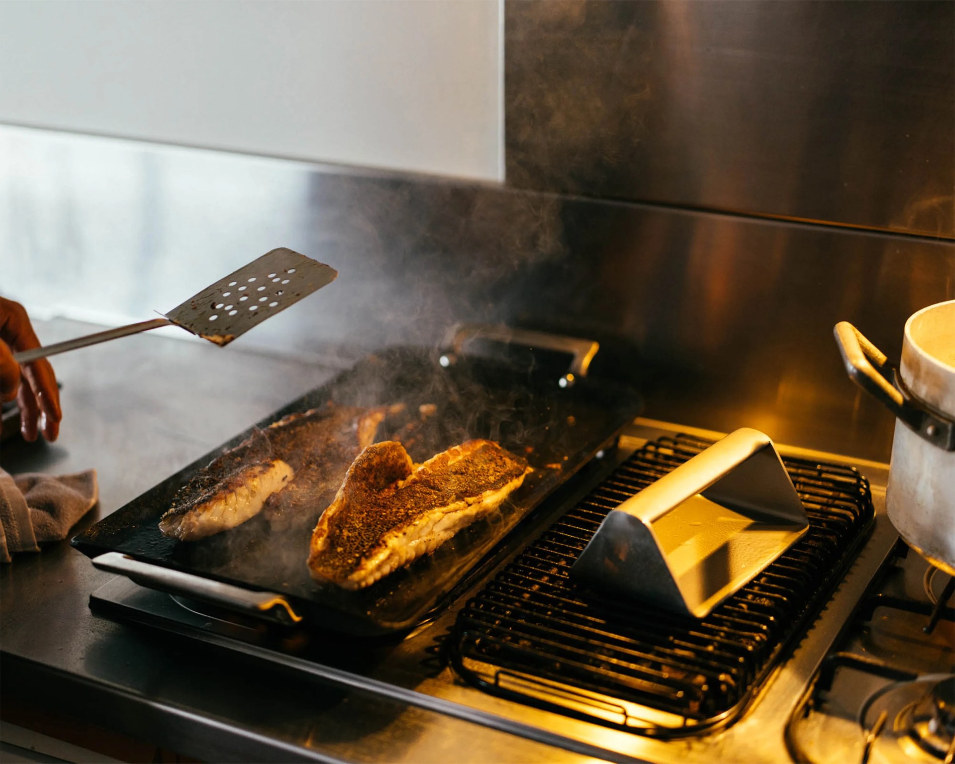 Two seasoned fish fillets cooking on a black griddle over a gas stove with a metal spatula in a hand.