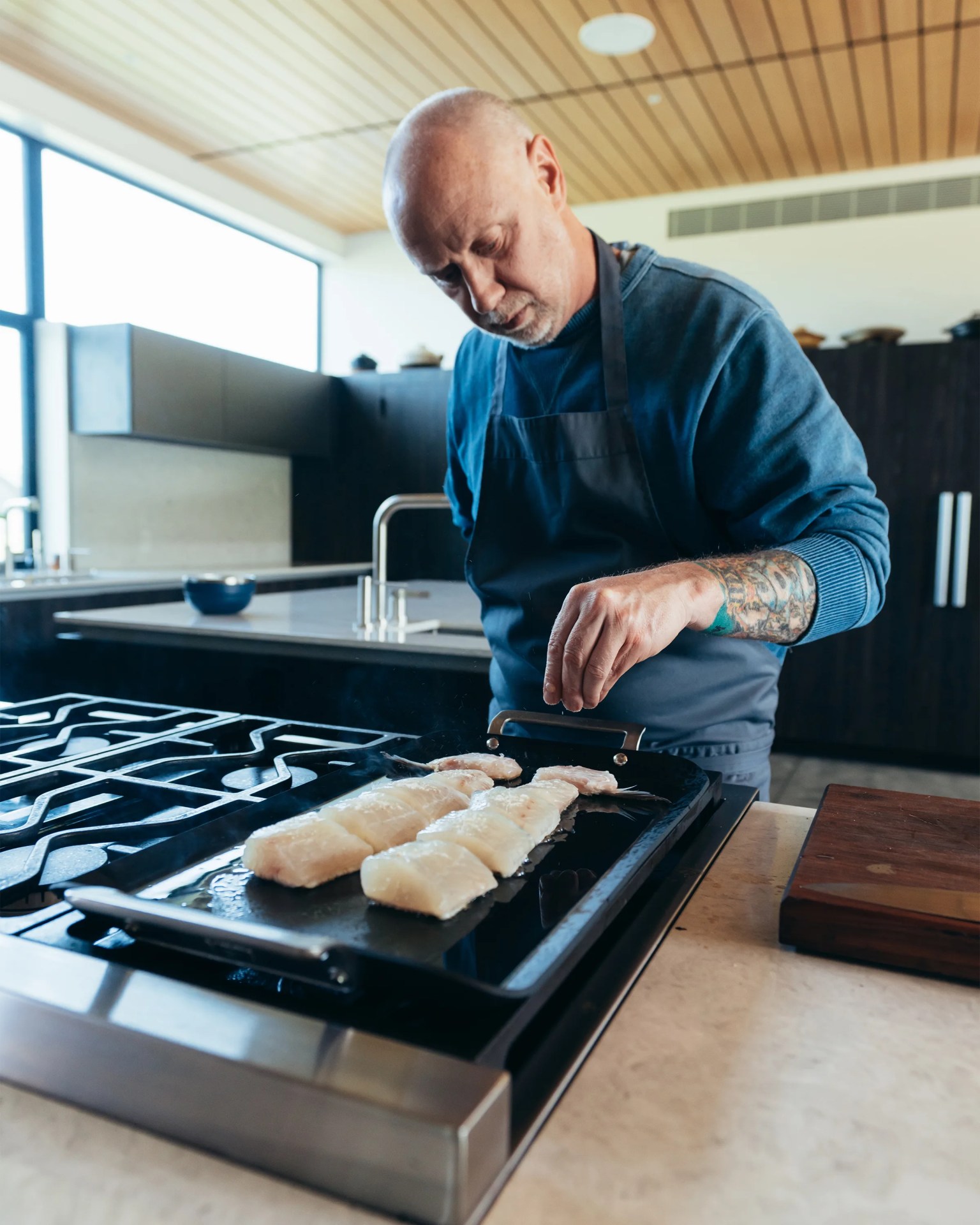 Man with tattooed forearm seasoning fish fillets on a black griddle in a modern kitchen.