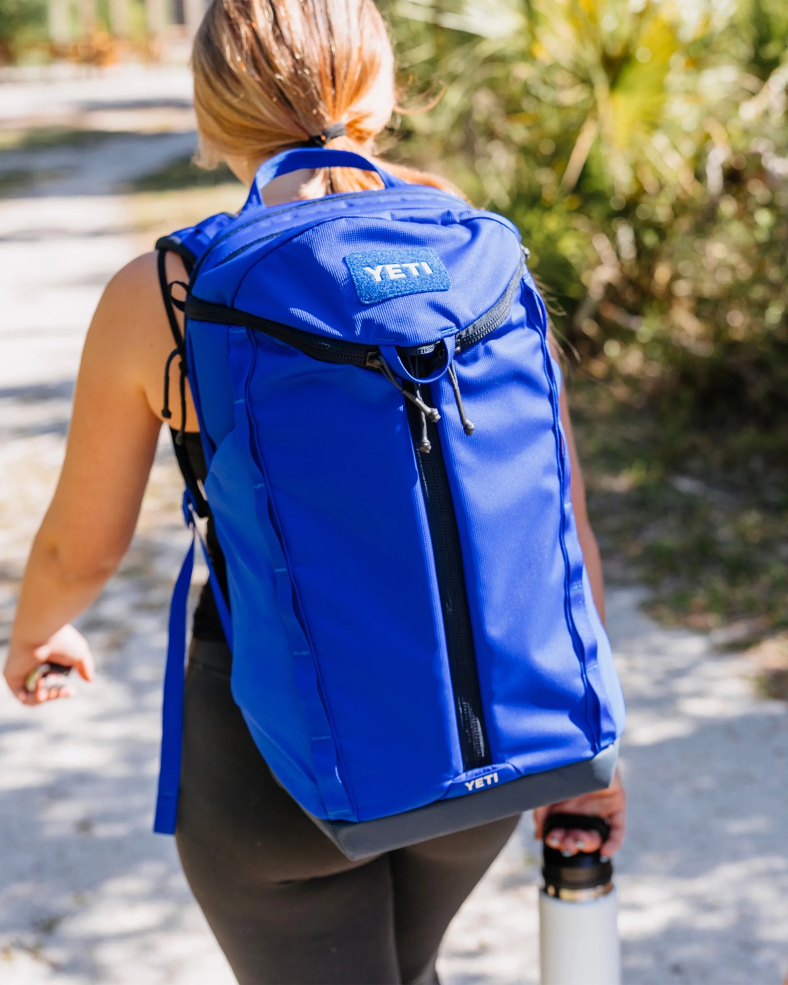 Bright blue YETI backpack with black zipper and handle worn by a person holding a white water bottle outdoors.