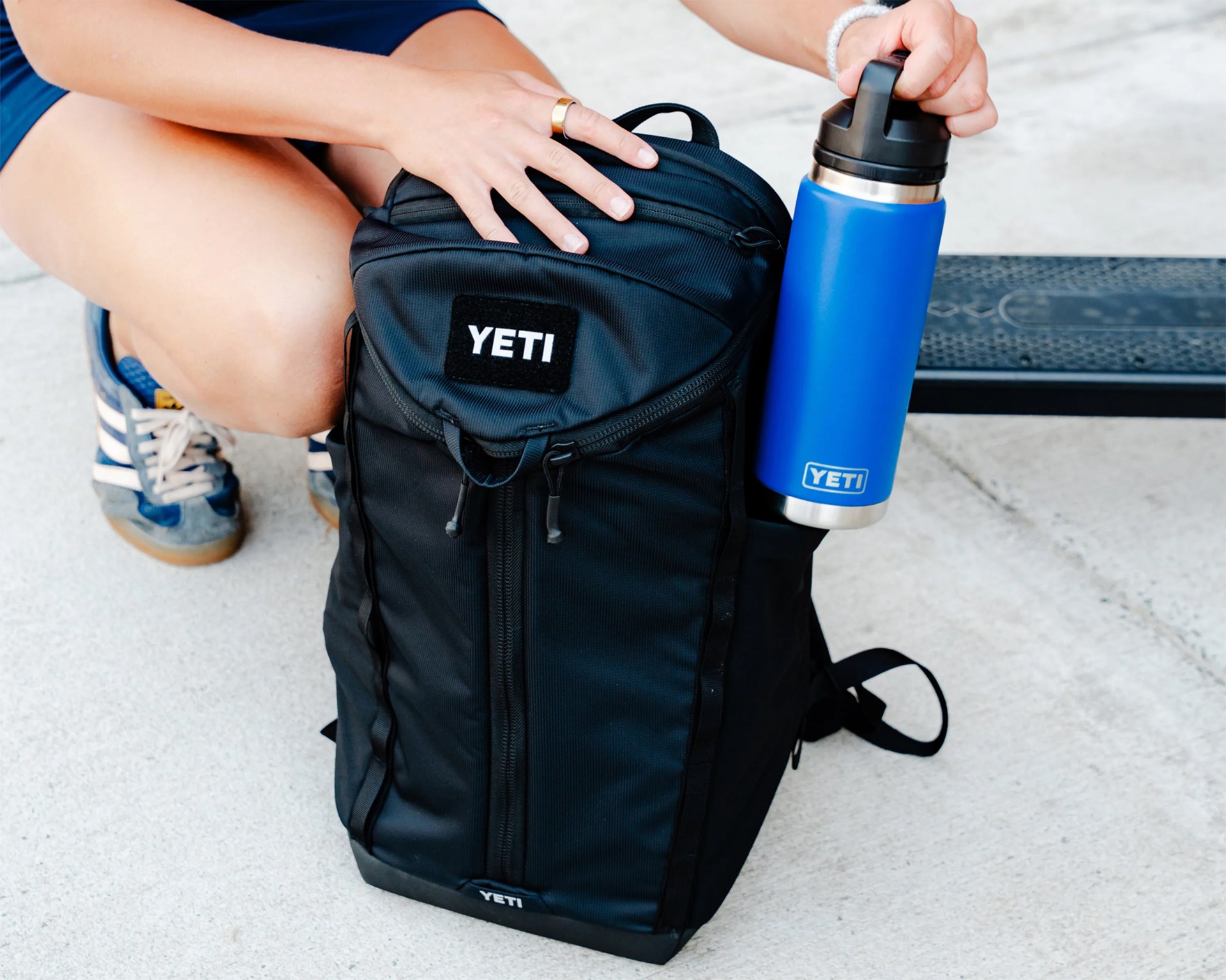 Black YETI backpack with a blue YETI insulated bottle held by a person wearing sneakers.