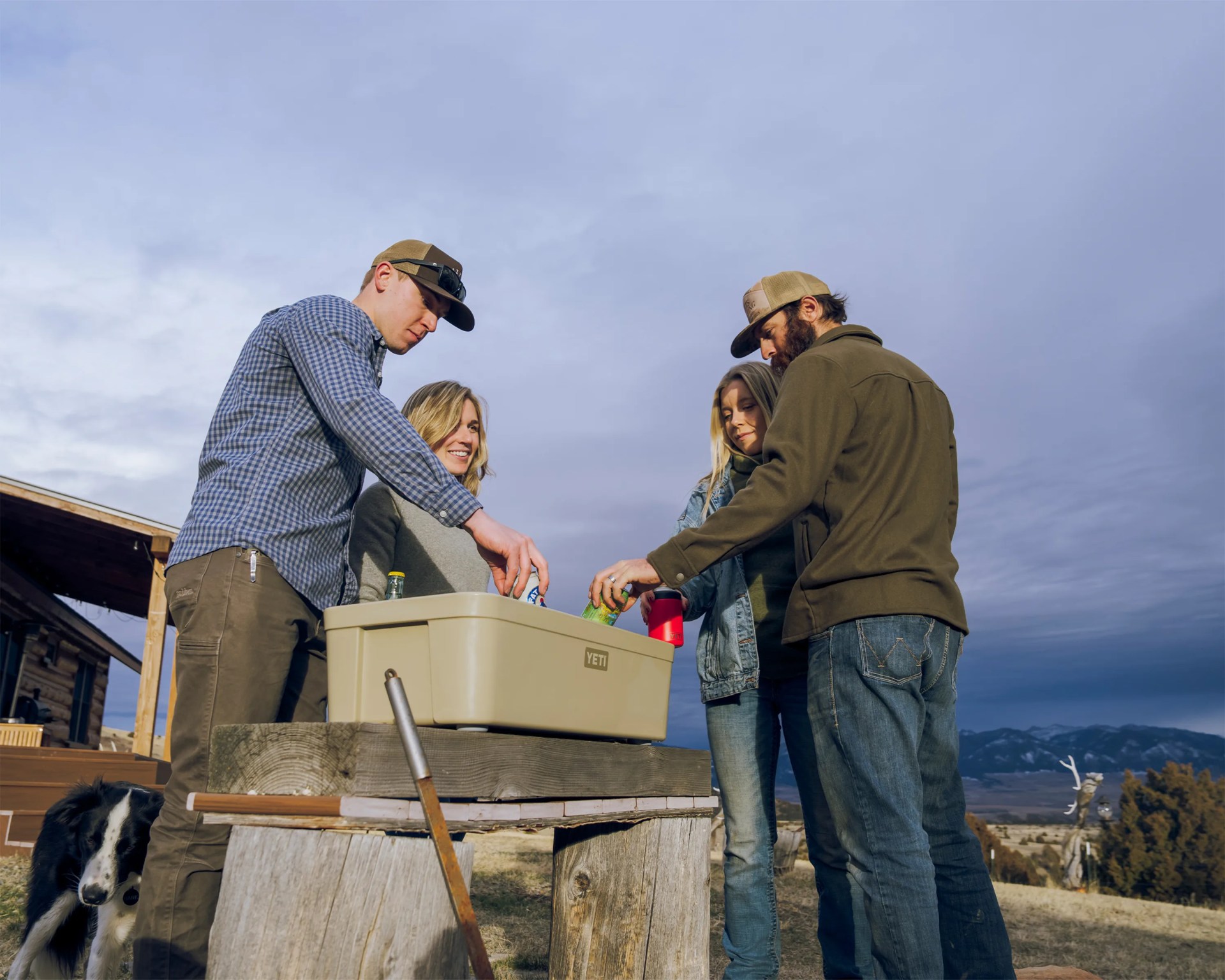 Four people wearing casual outdoor clothing and caps stand around a beige YETI cooler on a wooden table outdoors.