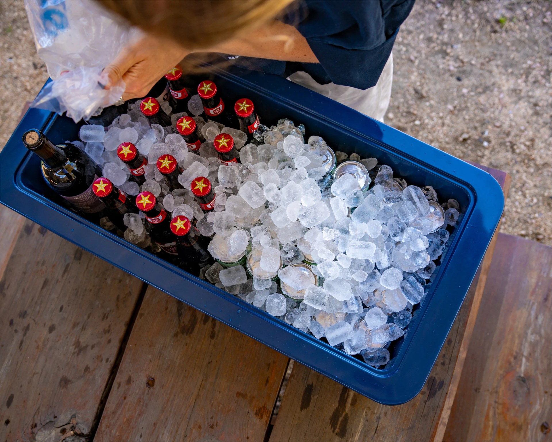 Blue cooler filled with ice, red-capped beer bottles, and a wine bottle on a wooden surface.