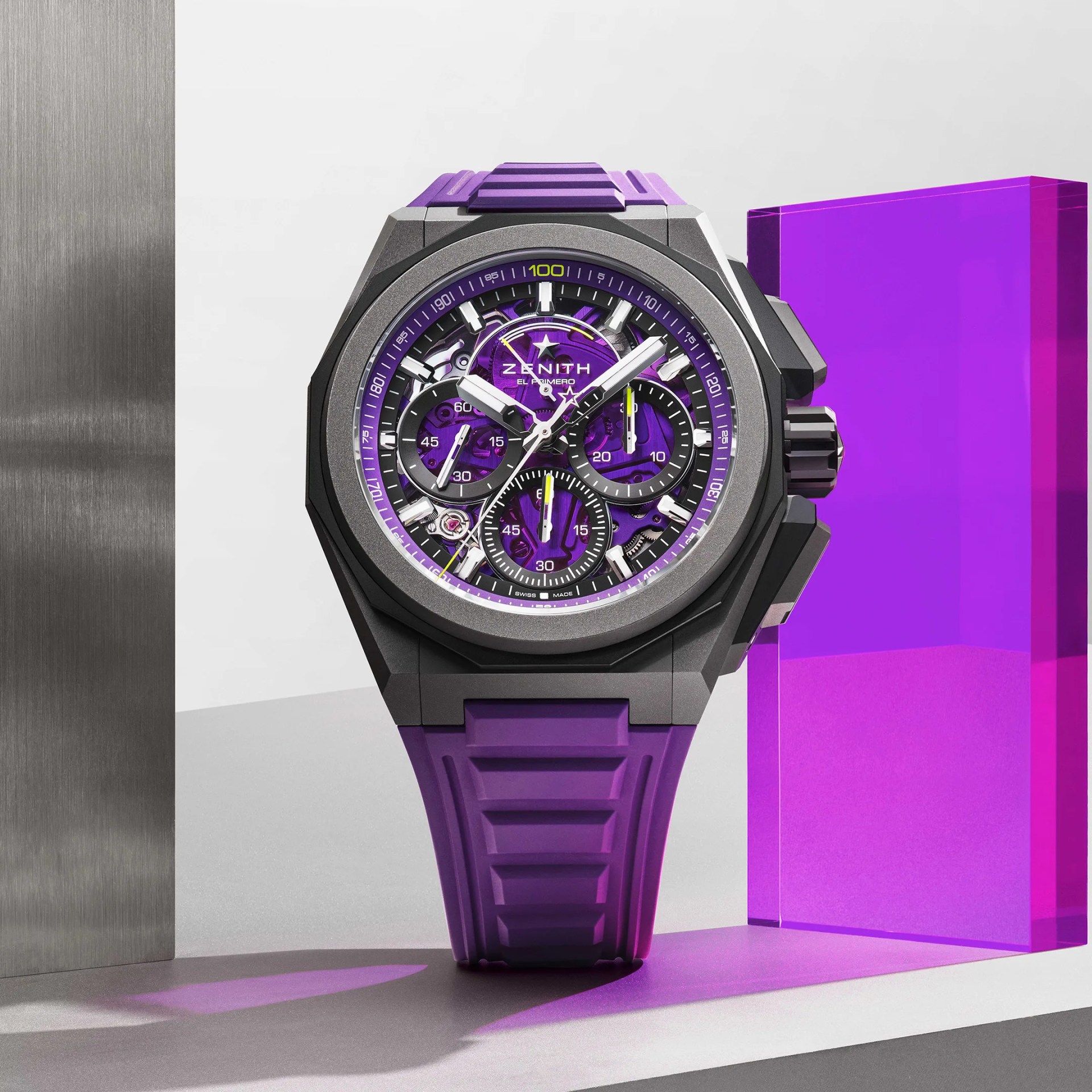 Black and gray Zenith El Primero watch with purple dial and strap, displayed with purple acrylic block.