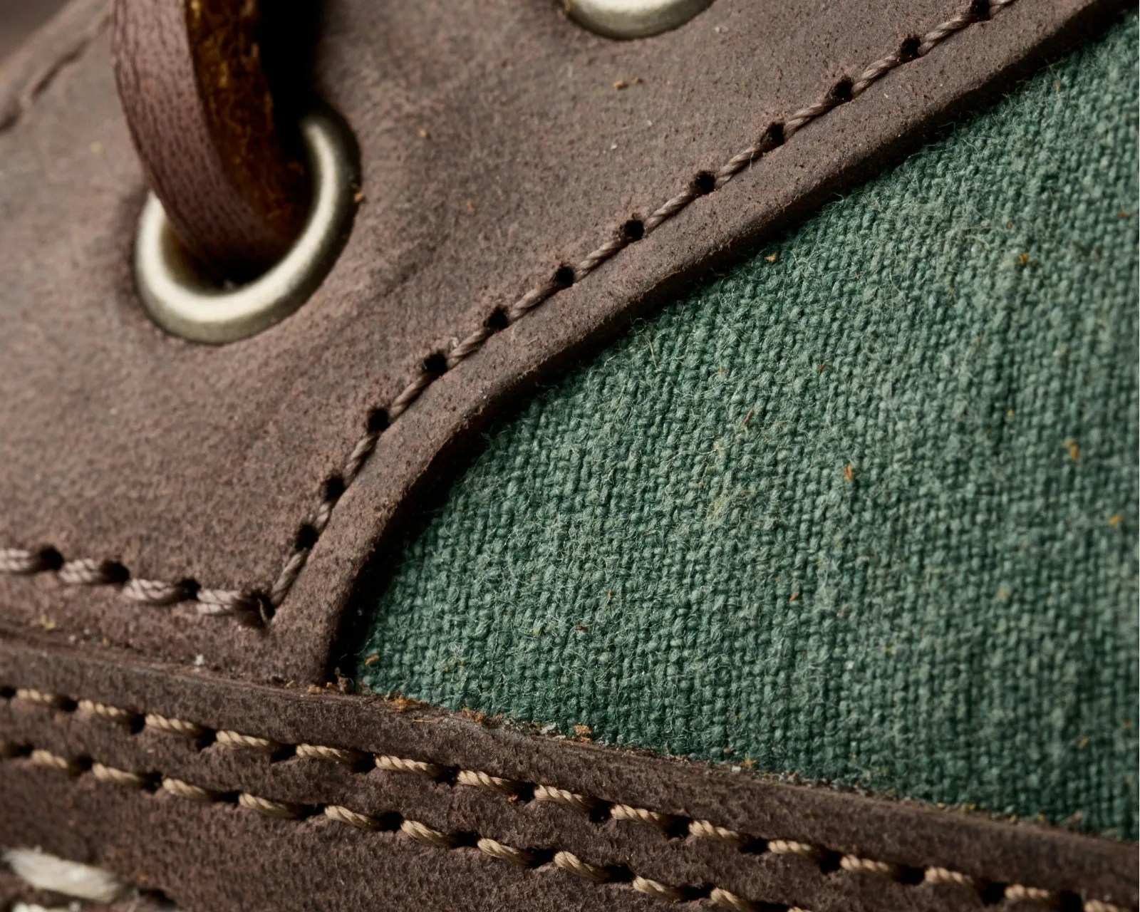 Close-up of brown suede and green woven fabric with detailed stitching and a metal eyelet on a shoe.