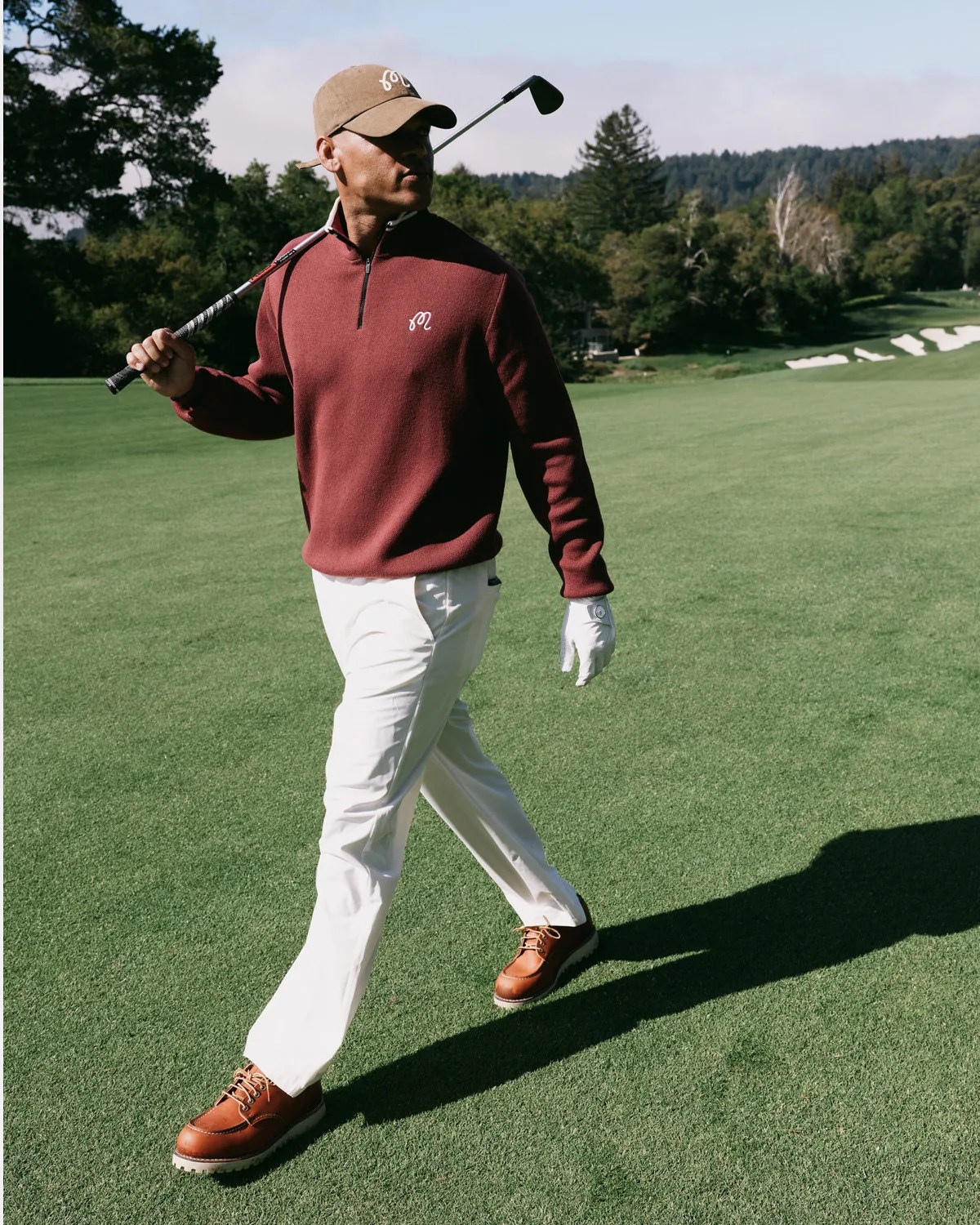 Person walking on a golf course wearing a maroon quarter-zip sweater, white pants, brown shoes, and a beige cap, holding a golf club.