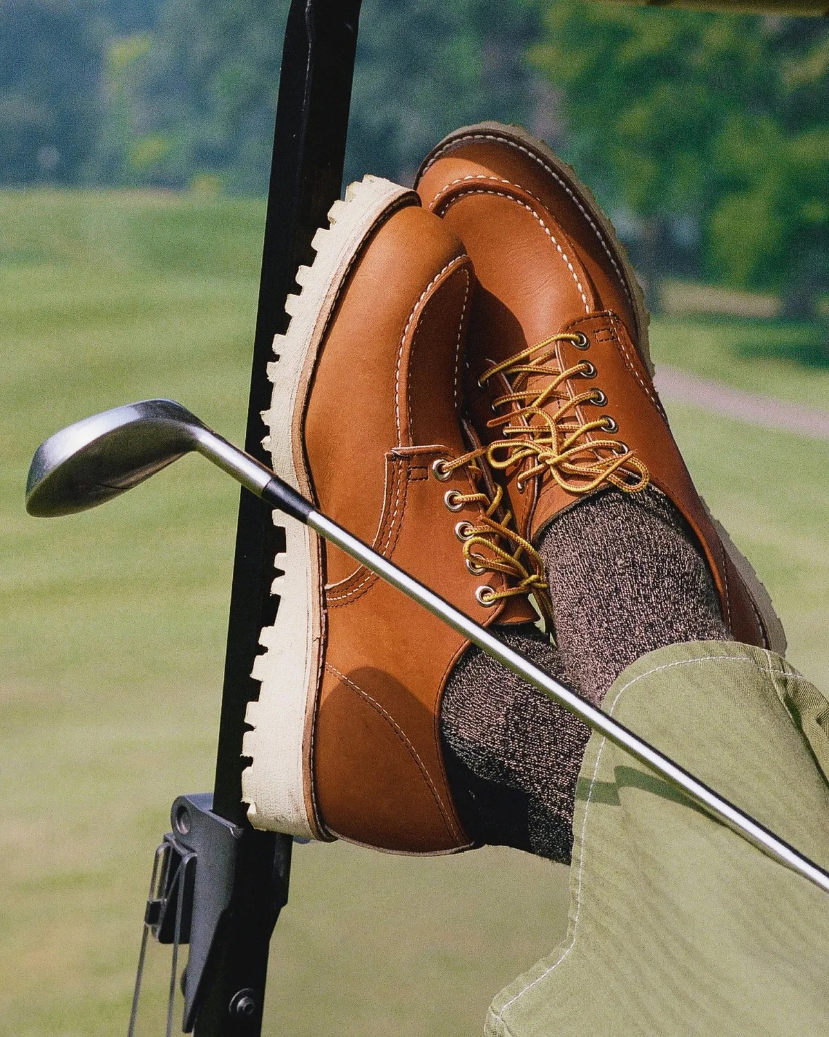 Brown leather lace-up shoes with beige soles worn with dark socks and green pants, resting on a golf cart with a golf club.