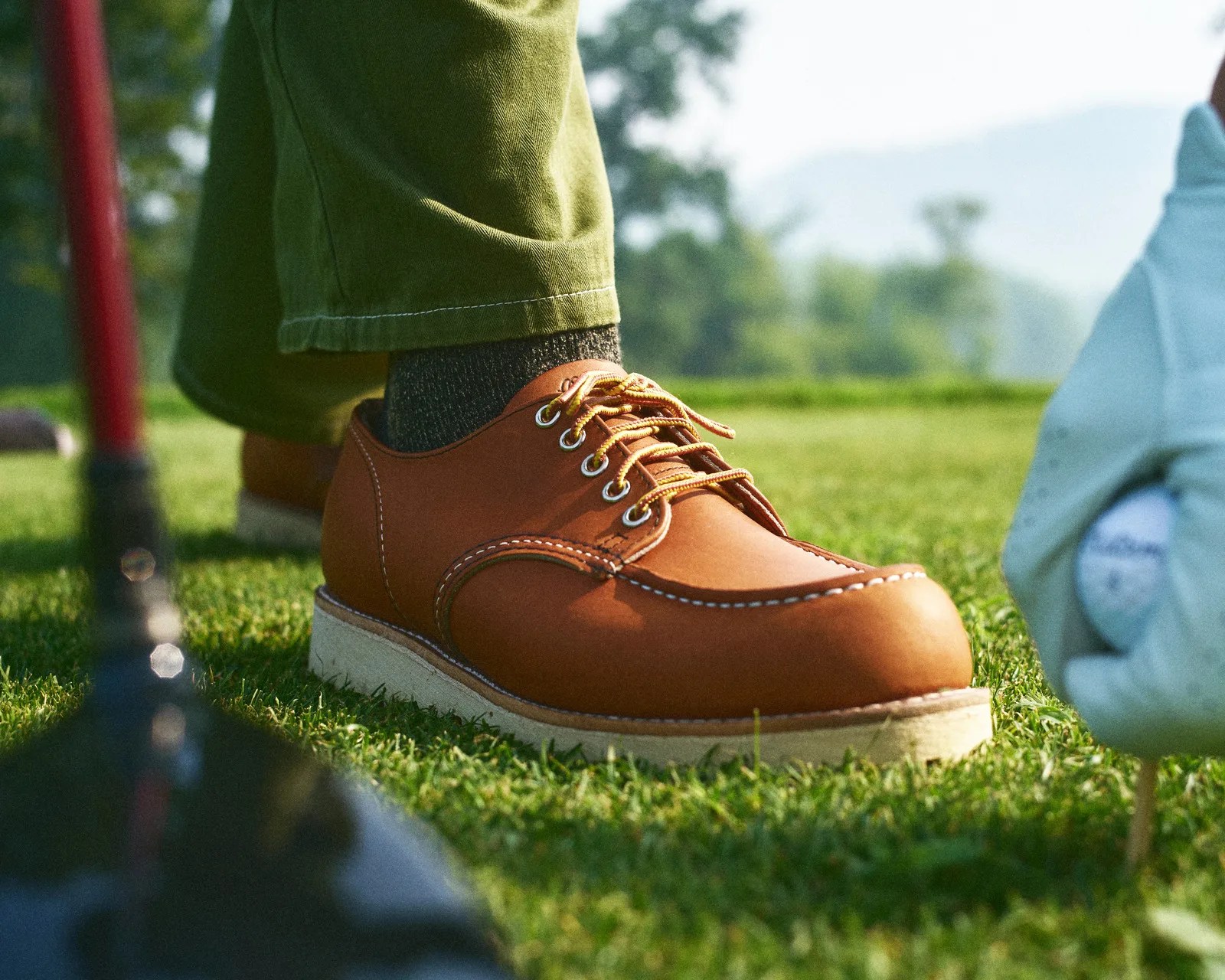 Brown leather golf shoe with yellow laces worn with green pants on grass near a golf ball in a white cloth bag.