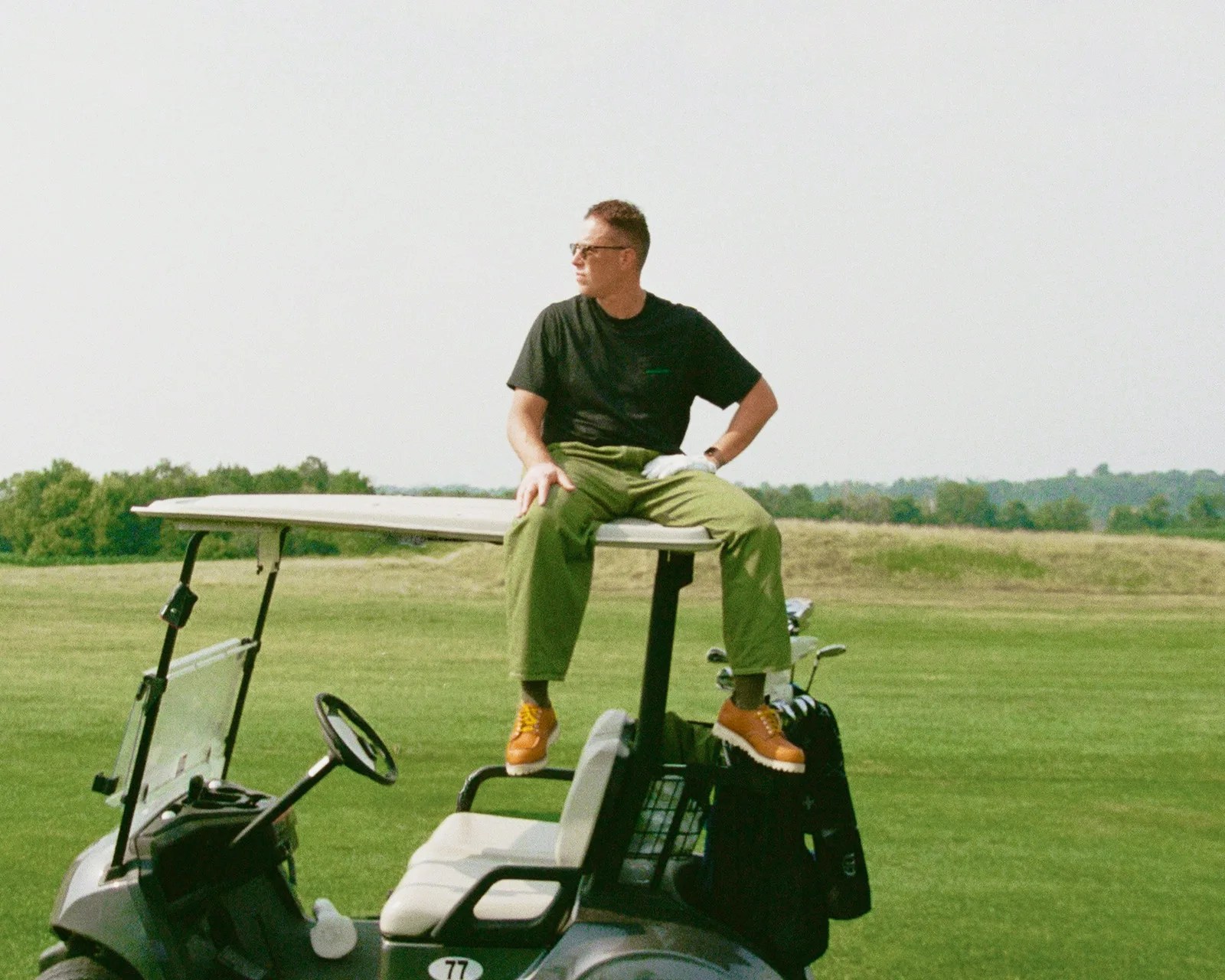 Man in black t-shirt, green pants, and brown shoes sitting on roof of a golf cart on a grassy field.