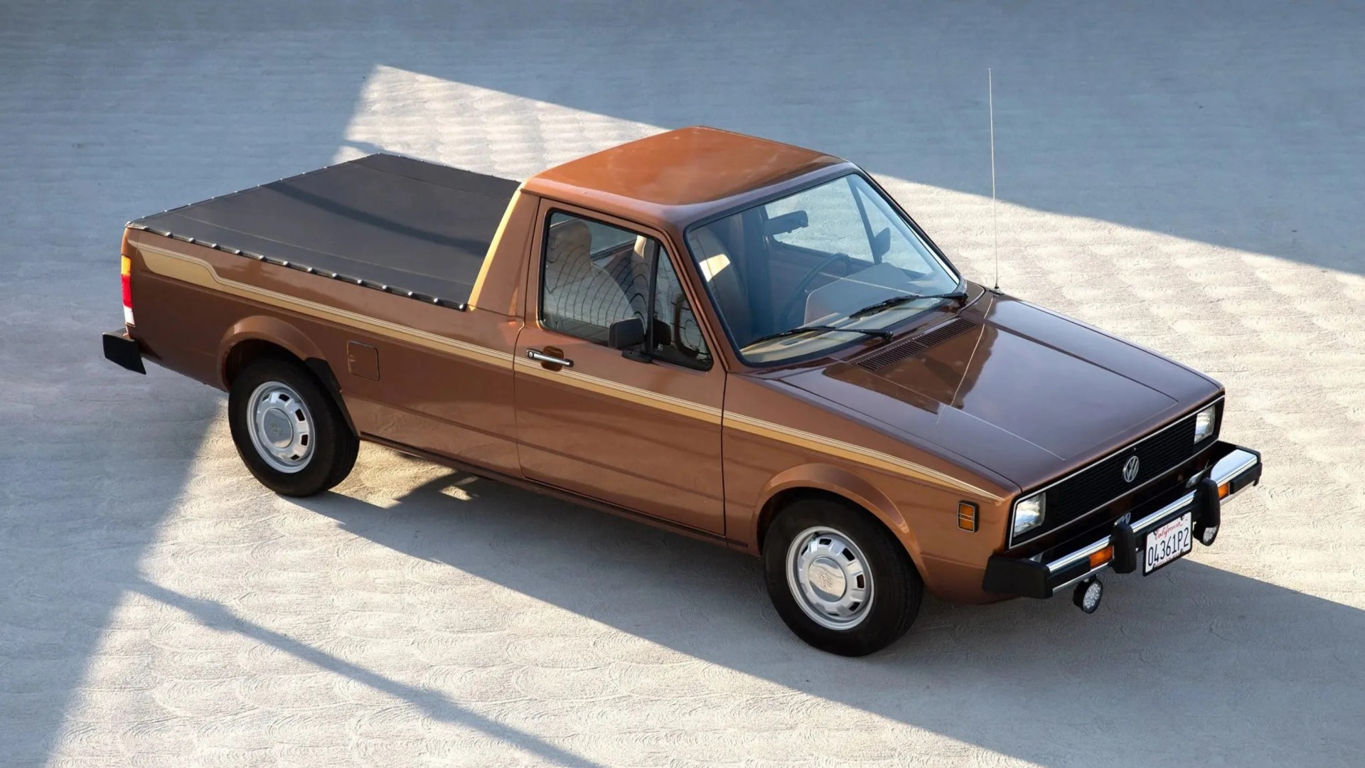 Brown vintage Volkswagen pickup truck with black tonneau cover parked on a light textured surface.