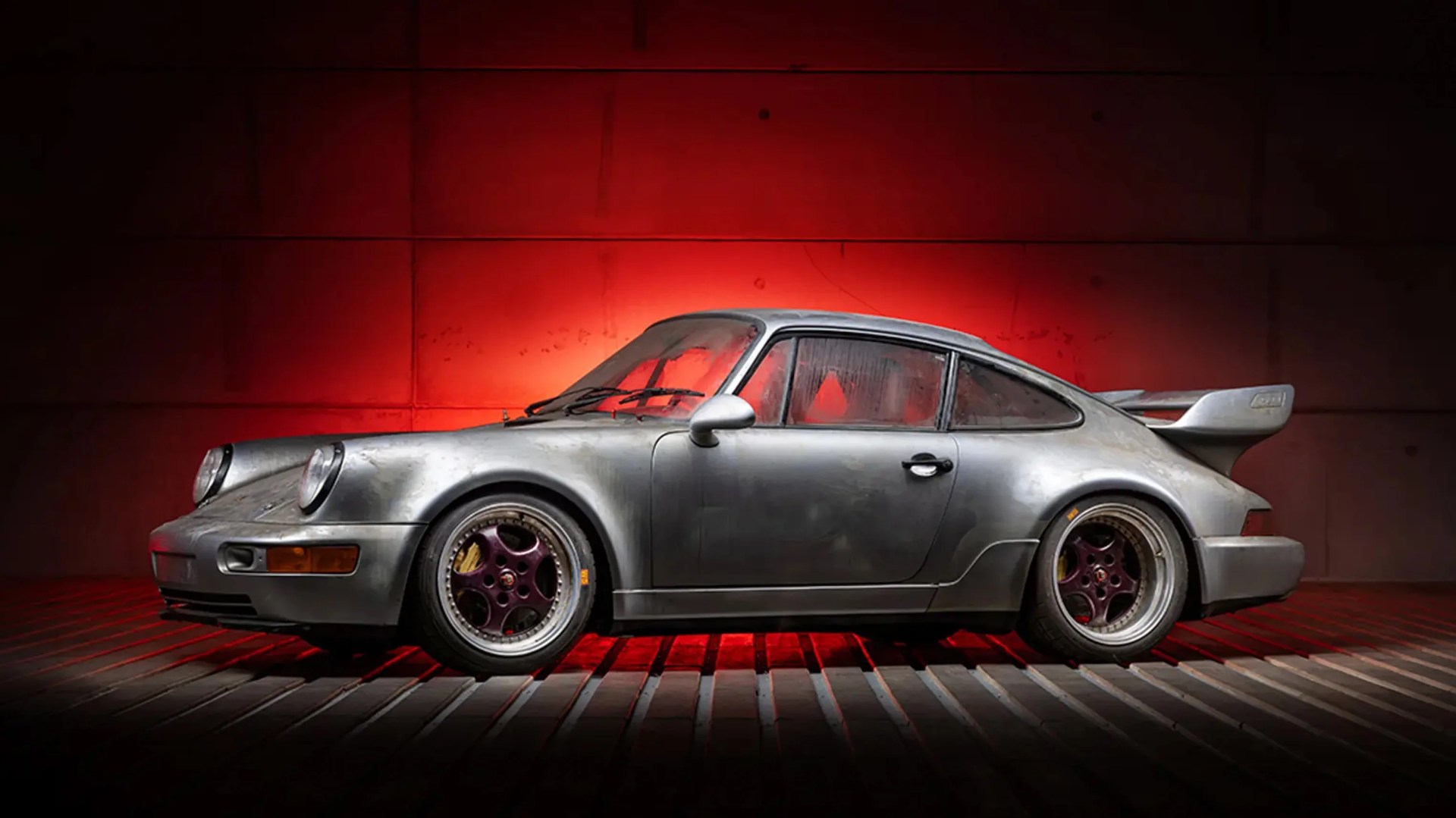 Silver vintage Porsche 911 with pop-up headlights and rear spoiler against a red-lit wall.
