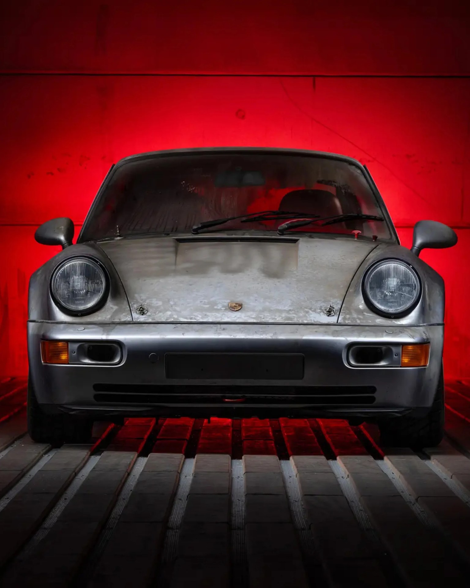 Front view of a dusty silver Porsche sports car with round headlights against a red background.