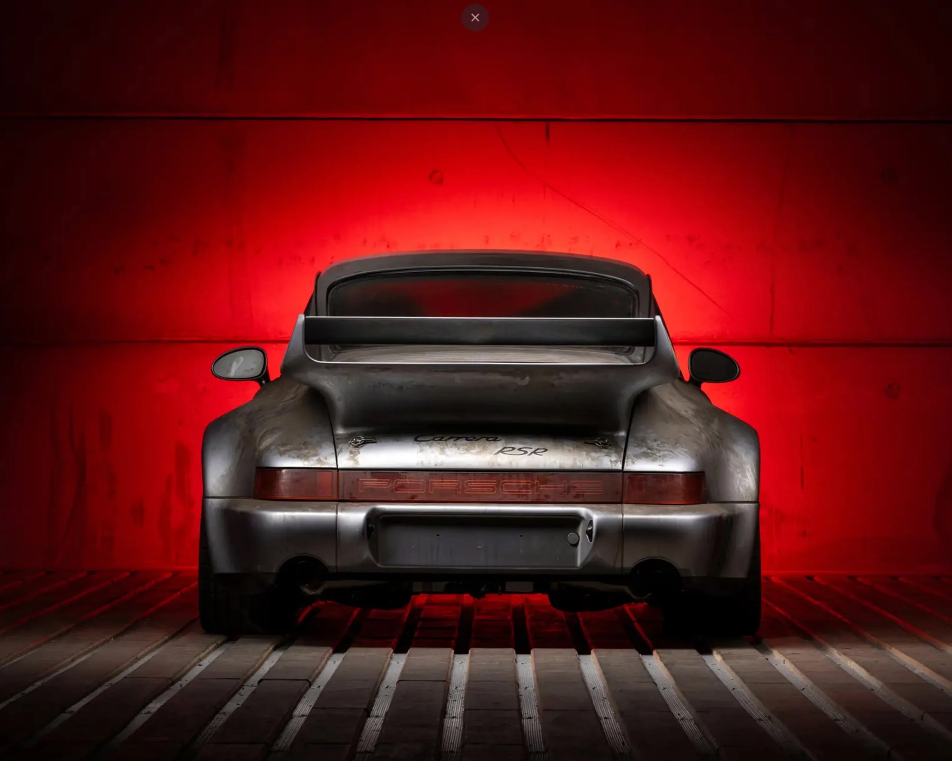 Rear view of a weathered silver Porsche Carrera RS with a large rear spoiler against a red background.
