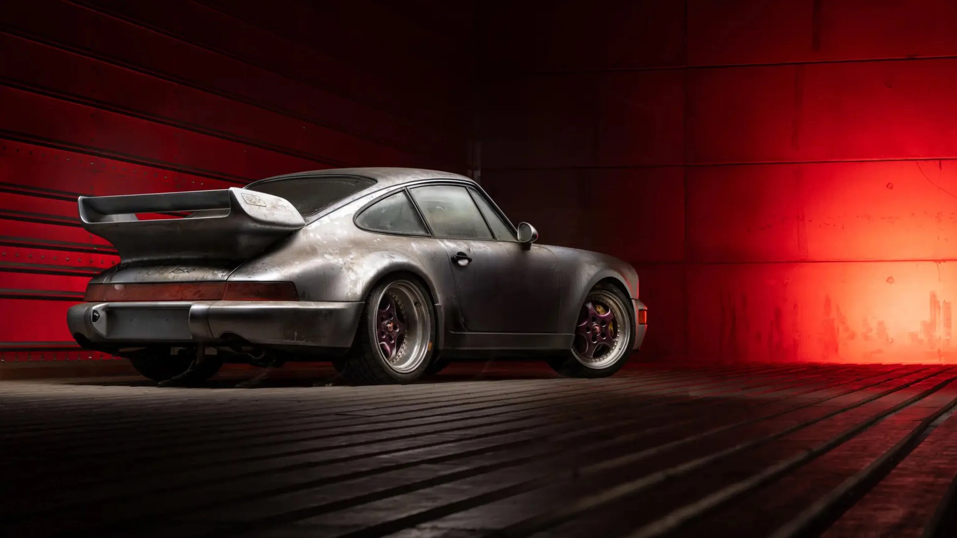 Silver vintage Porsche 911 with large rear spoiler and purple rims in a dimly lit red room.
