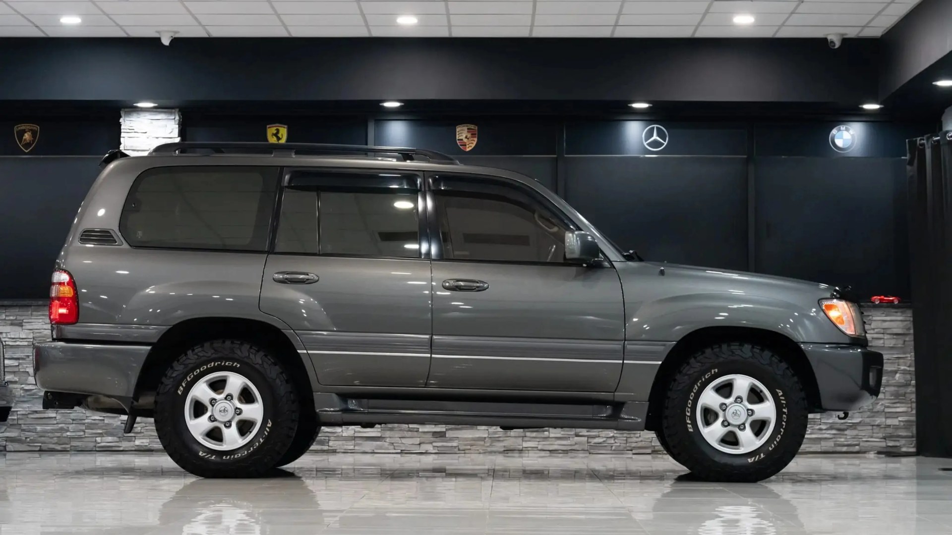 Gray Toyota Land Cruiser SUV with BFGoodrich tires parked indoors against a dark wall with luxury car logos.