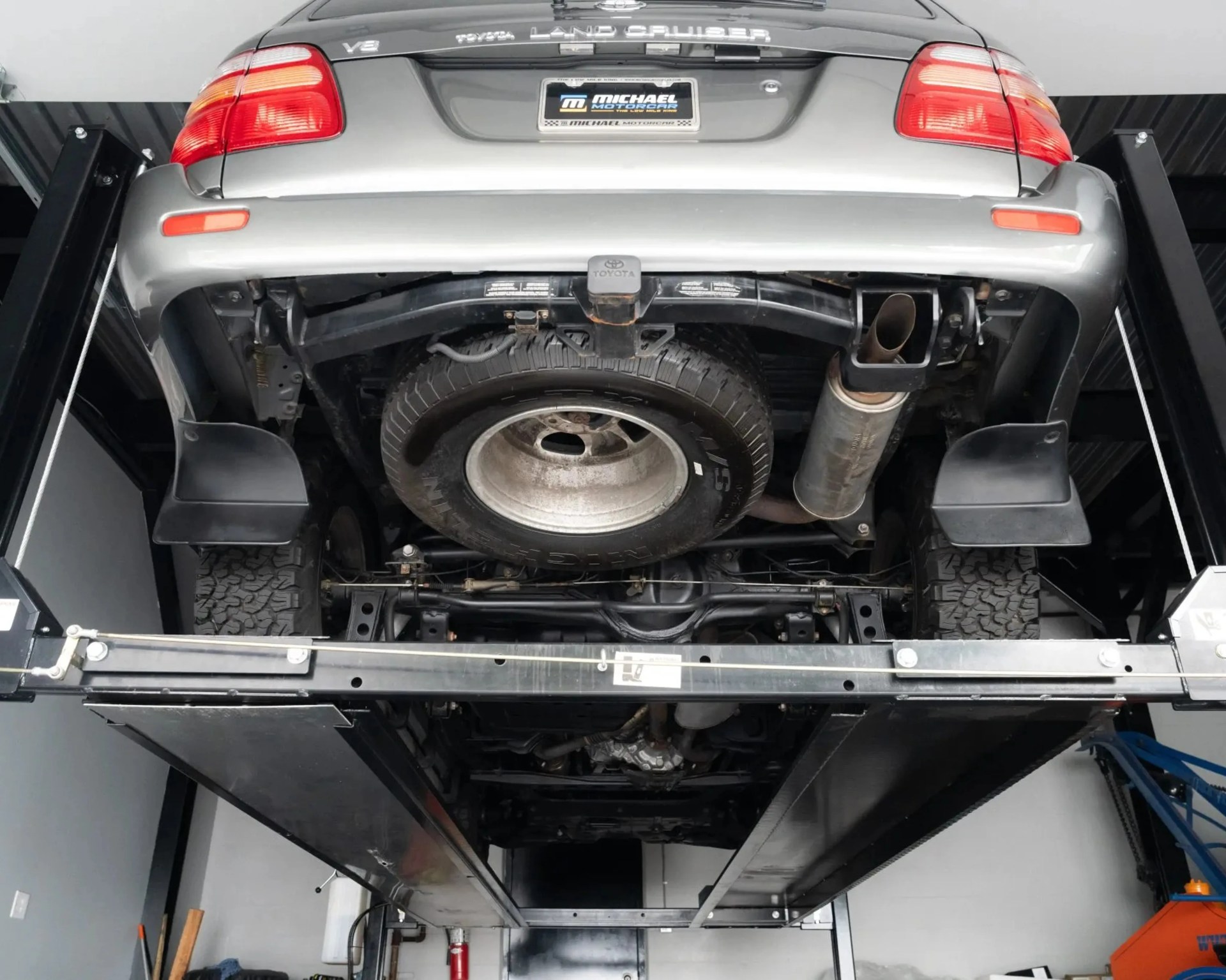 Underside view of a silver Toyota Land Cruiser with spare tire mounted under the rear bumper on a car lift in a garage.