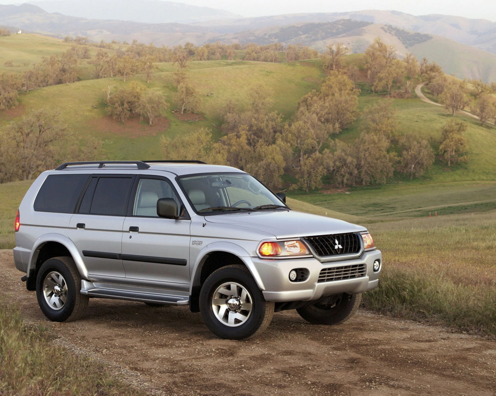 Silver Mitsubishi Montero Sport SUV parked on a dirt path with grassy hills and trees in the background.