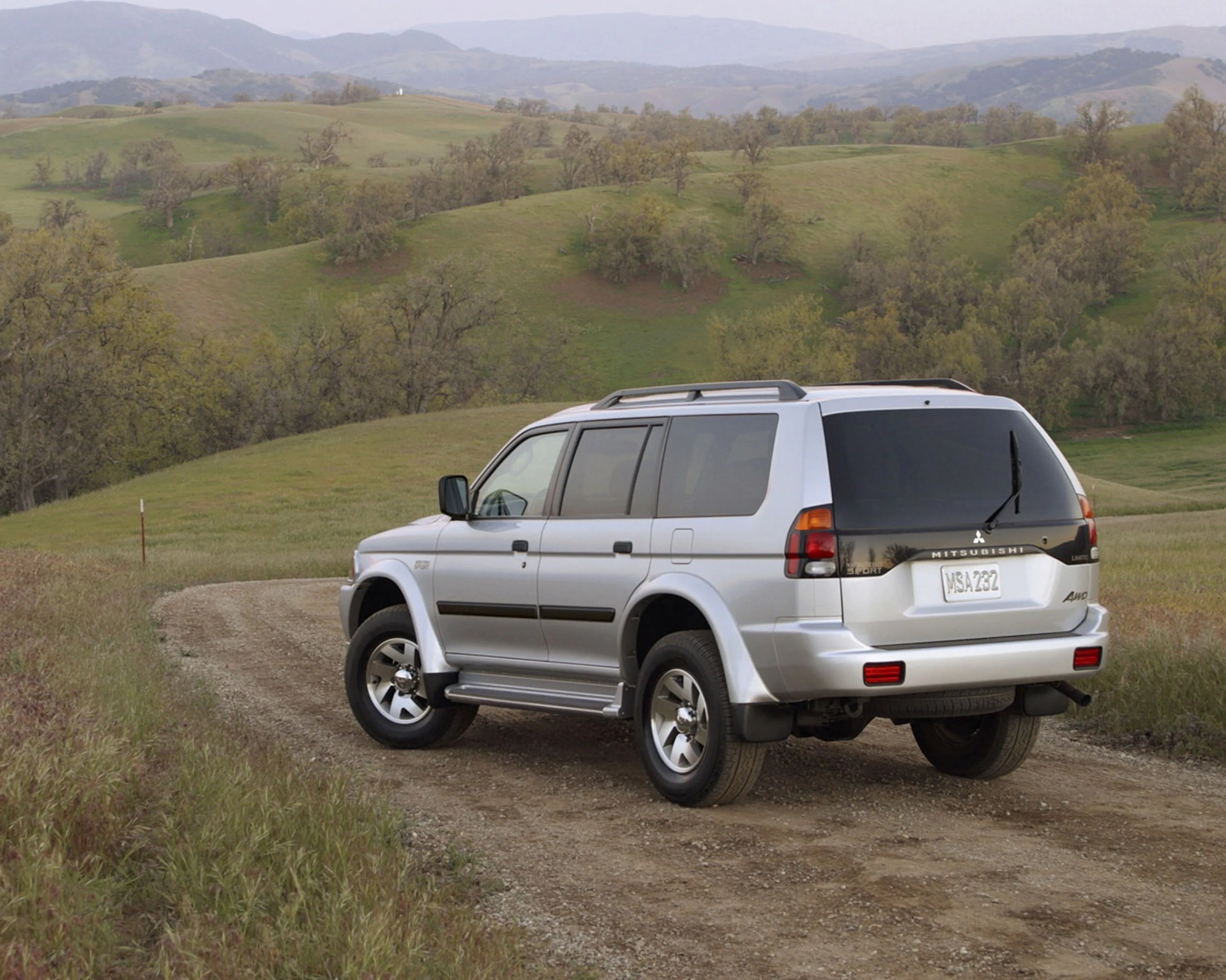 Silver Mitsubishi SUV parked on a dirt road with green hills and trees in the background.