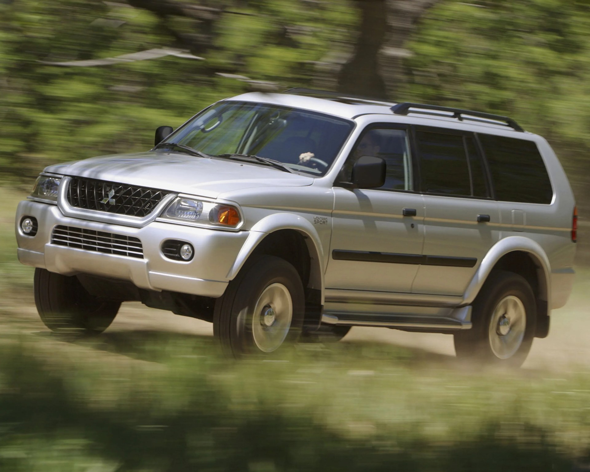 Silver Mitsubishi Pajero Sport SUV driving on a dirt road with blurred green foliage background.