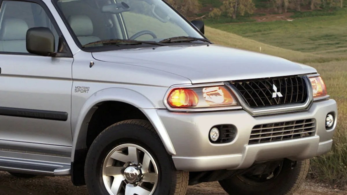 Silver Mitsubishi Montero Sport SUV front side with headlights on, parked on a dirt path near grass.