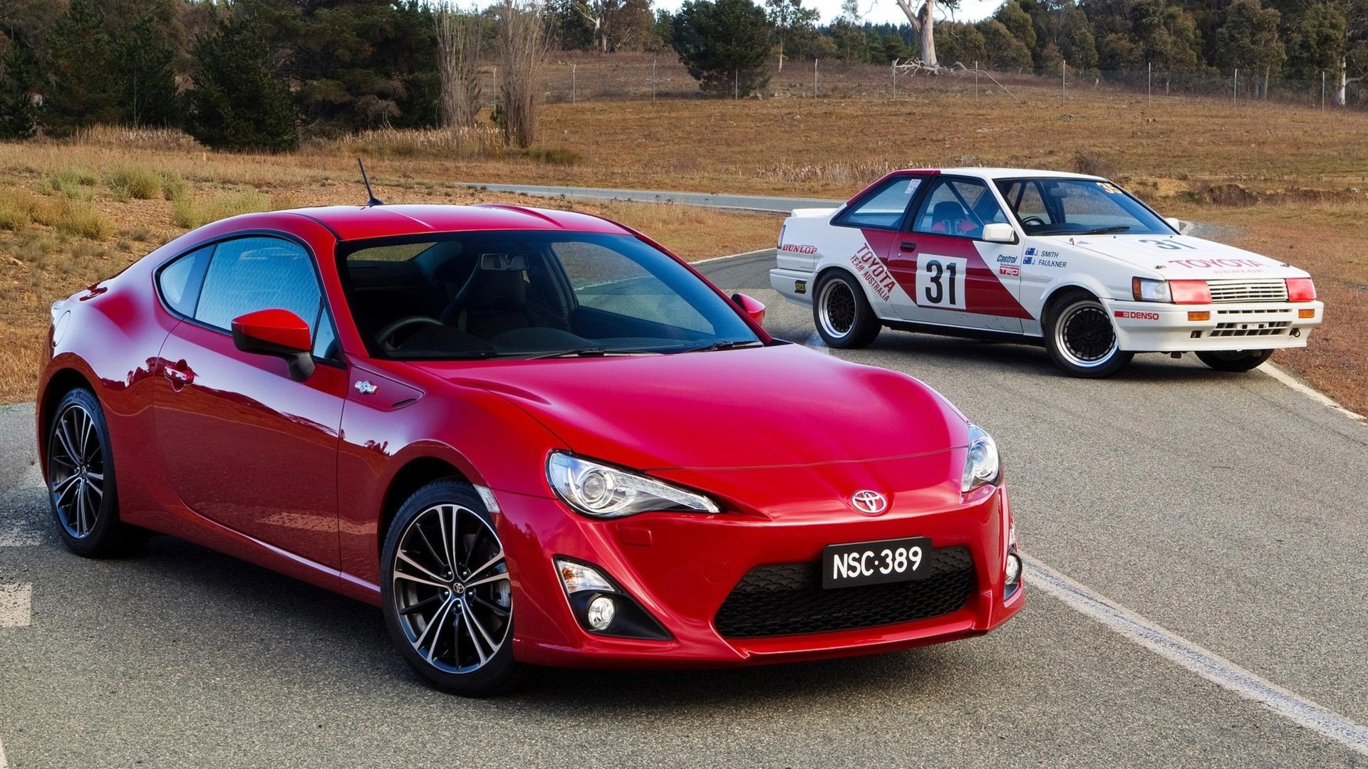 Red modern Toyota sports car in front of a white vintage Toyota race car on a rural road.