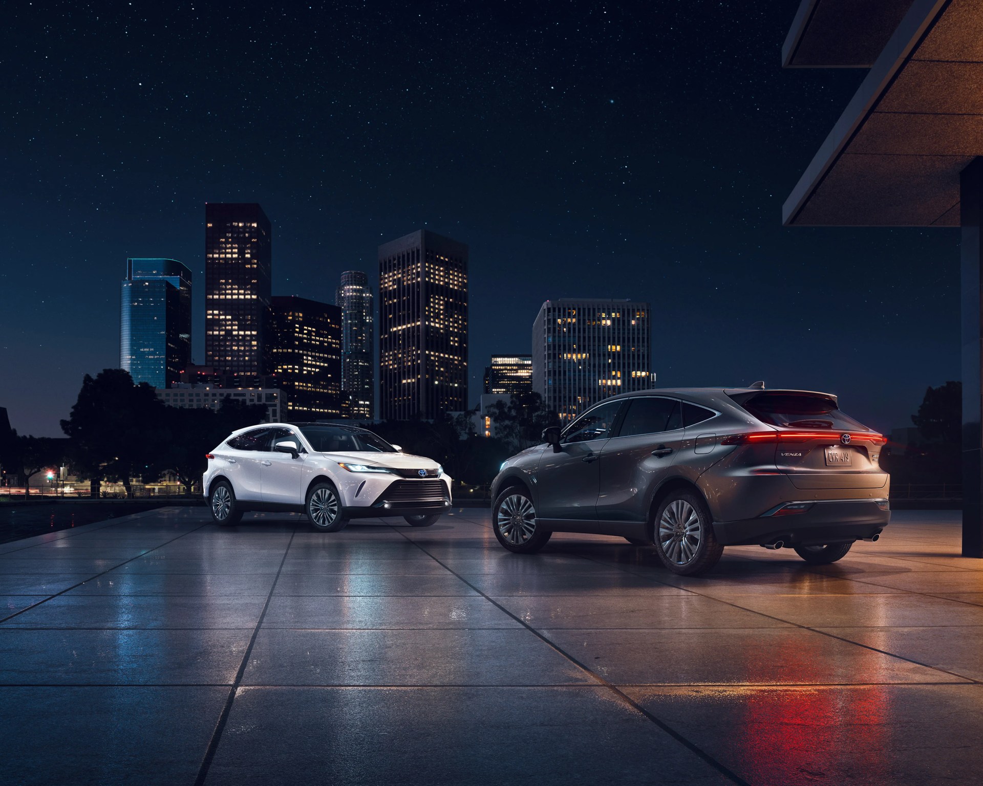 Two Toyota Venza SUVs, one white and one gray, parked on a tiled surface with a city skyline and starry night sky in the background.