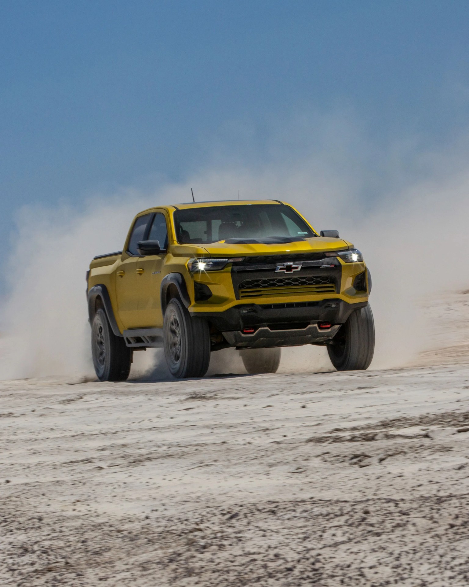 Yellow Chevrolet pickup truck driving on a dusty, sandy terrain with headlights on.
