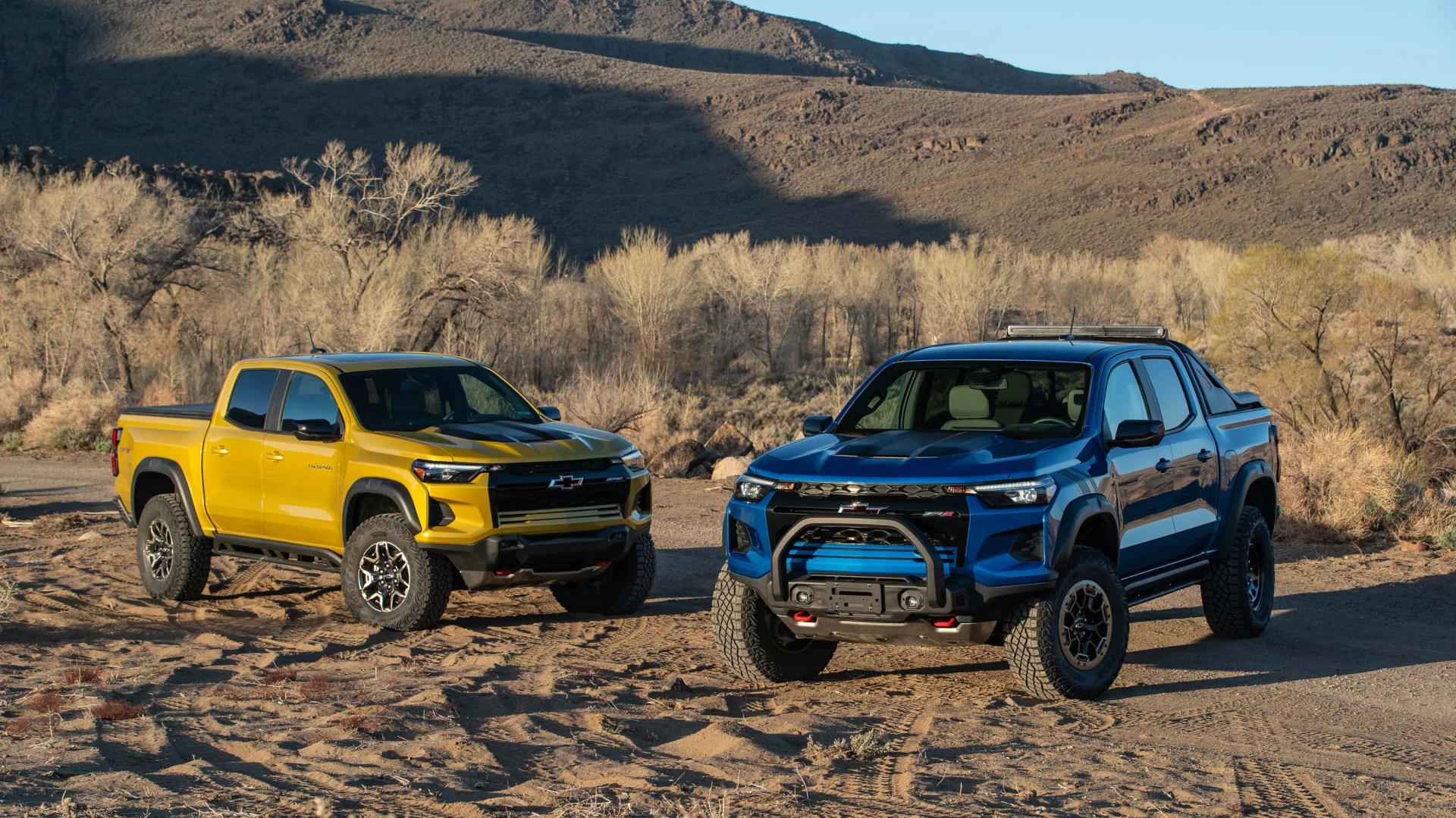 Two Chevrolet pickup trucks, one yellow and one blue, parked on a dirt area with dry trees and hills in the background.